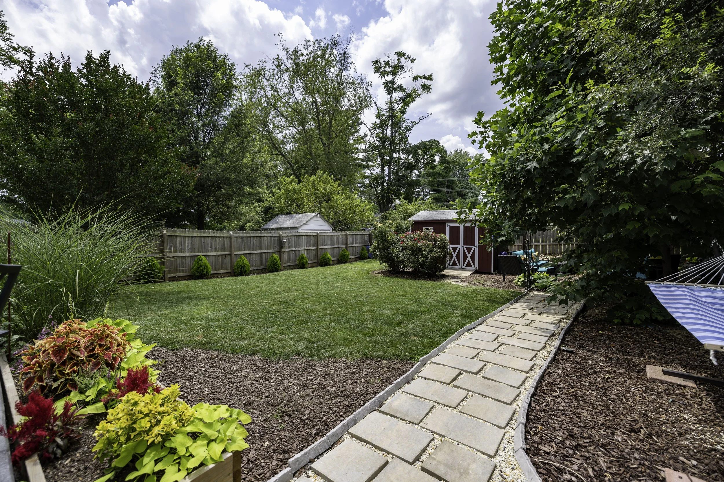 A backyard garden with a stone pathway, a grassy lawn, various plants and bushes, a wooden fence, a red garden shed, and trees under a partly cloudy sky.