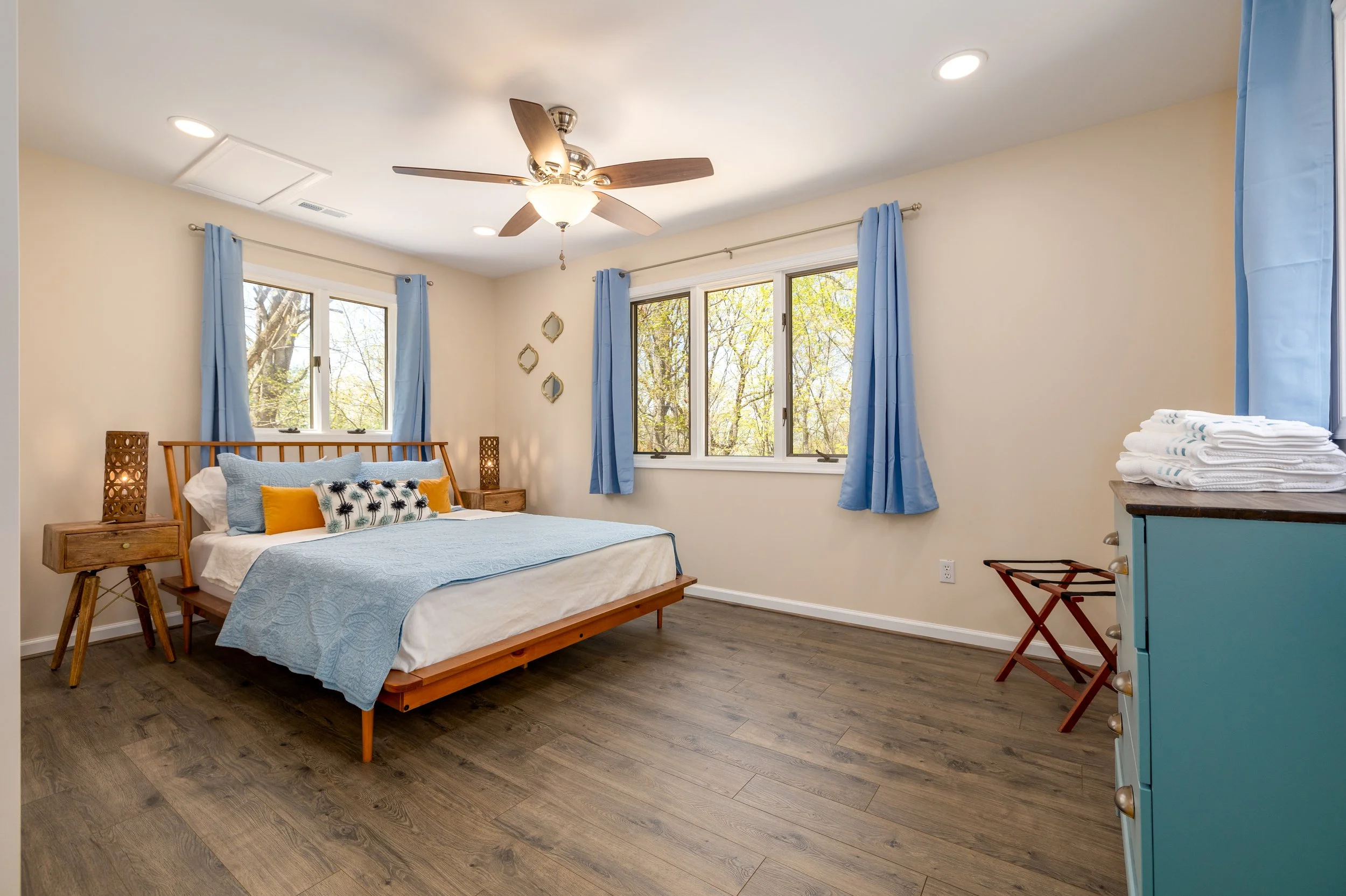 A bright bedroom with large windows, a wooden bed with a light blue quilt, decorative pillows, blue curtains, a ceiling fan with wooden blades, and a dresser with folded towels.