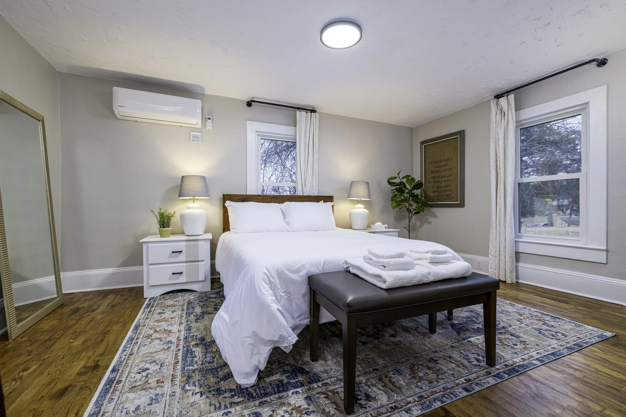 A cozy bedroom with a neatly made bed covered in white linens. Two bedside tables with lamps and plants, a bench with folded towels at the foot of the bed, and a large mirror. There are two windows with white curtains, hardwood floors, and an area rug. The ceiling holds a round light fixture and an air conditioning unit.