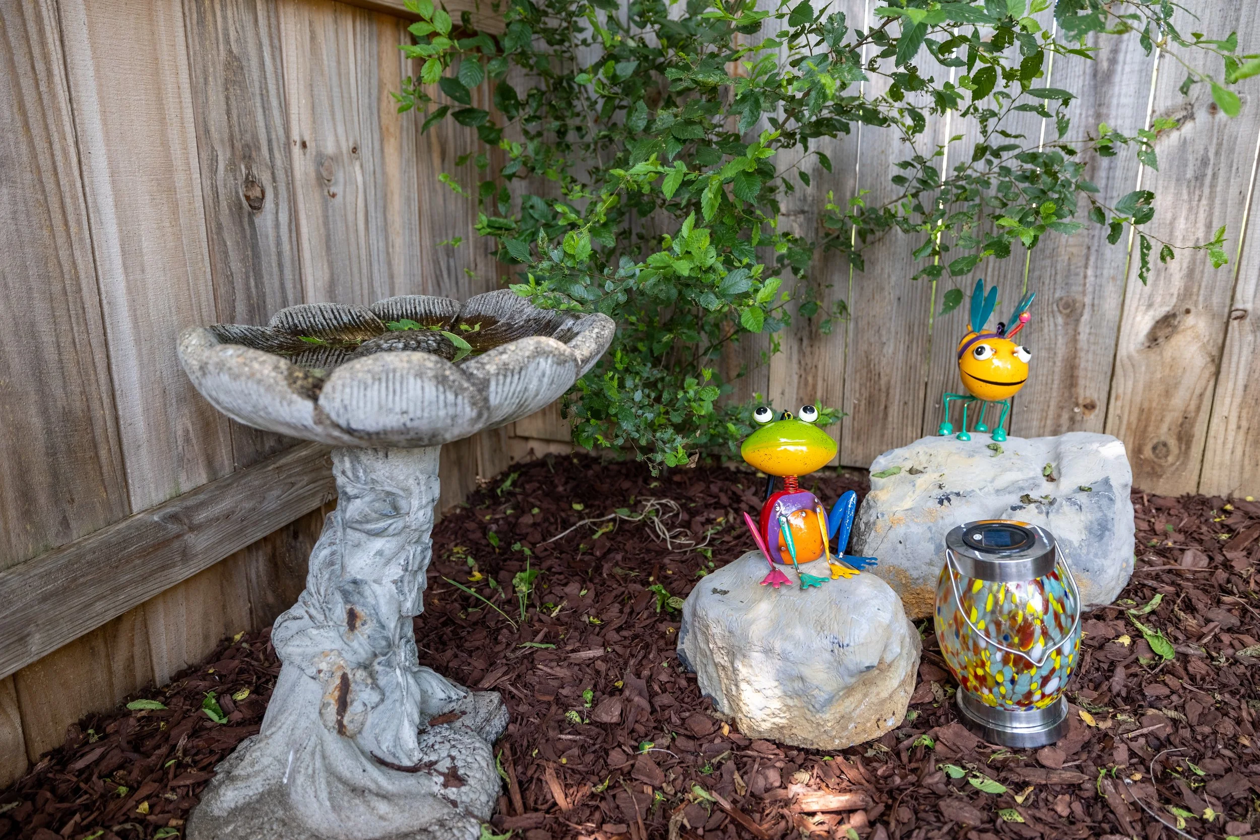 A backyard garden scene with a wooden fence, green plants, and colorful garden ornaments including a yellow smiling bug, a green frog, and a dragonfly. There is also a large decorative glass jar with a vibrant mosaic pattern.