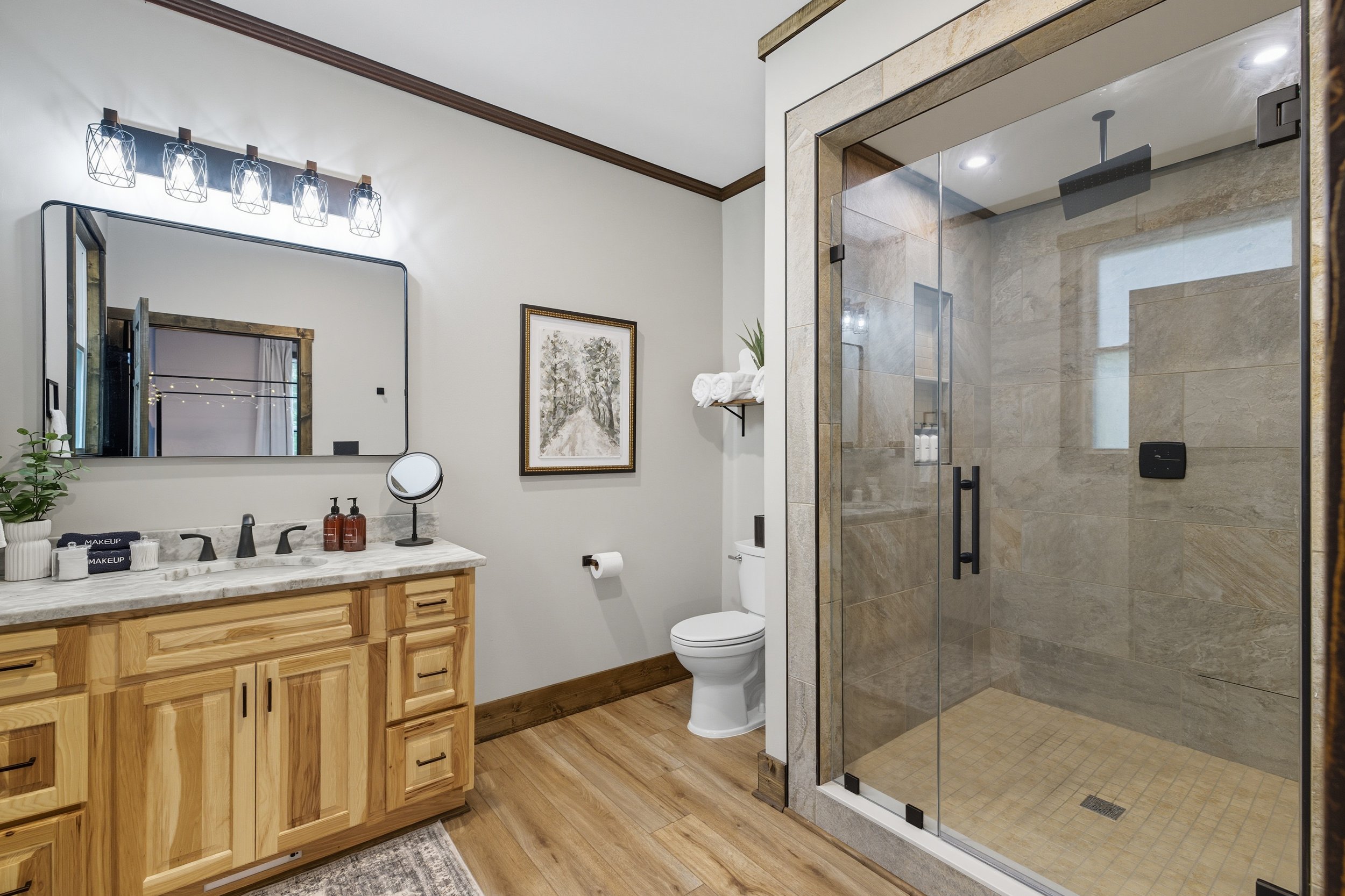 A modern bathroom with a wooden vanity, a large mirror, a white toilet, a glass-enclosed shower with beige tiles, a framed artwork on the wall, a shelf with rolled towels, and a small plant.