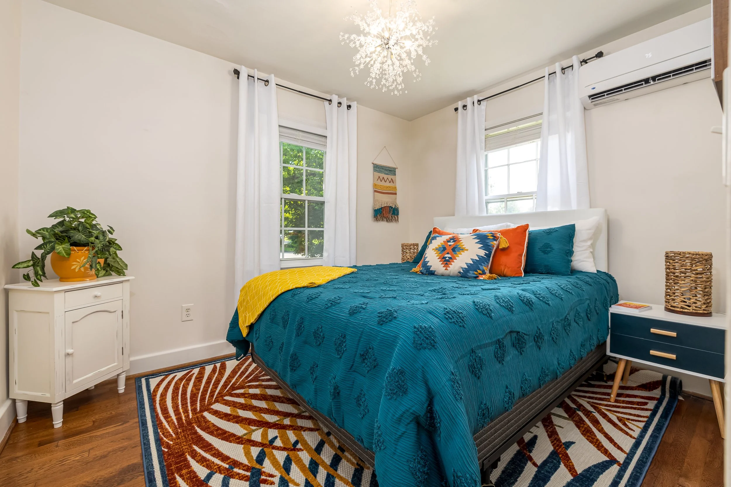A bedroom with a white bedspread, and colorful pillows, and a yellow throw. The room has white walls, two windows with white curtains, and a white nightstand with a potted plant. There is a patterned rug on a wooden floor and a decorative wall hangin