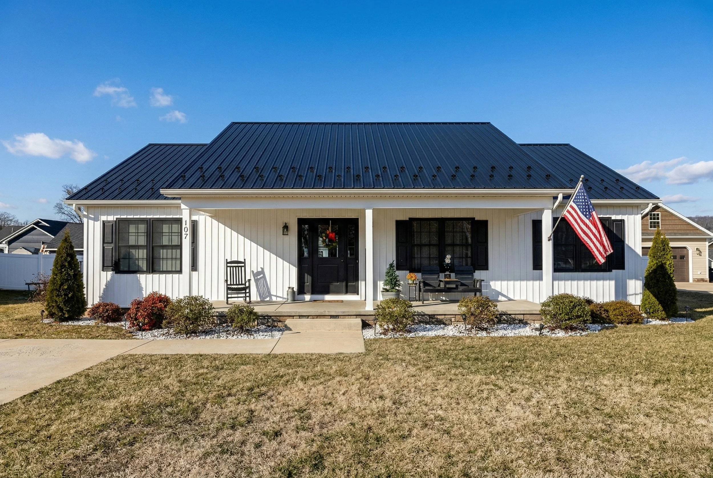 Front view of a white house with black shutters, a black metal roof, front porch, American flag on a pole, and landscaping including bushes and a small tree, under a blue sky with scattered clouds.