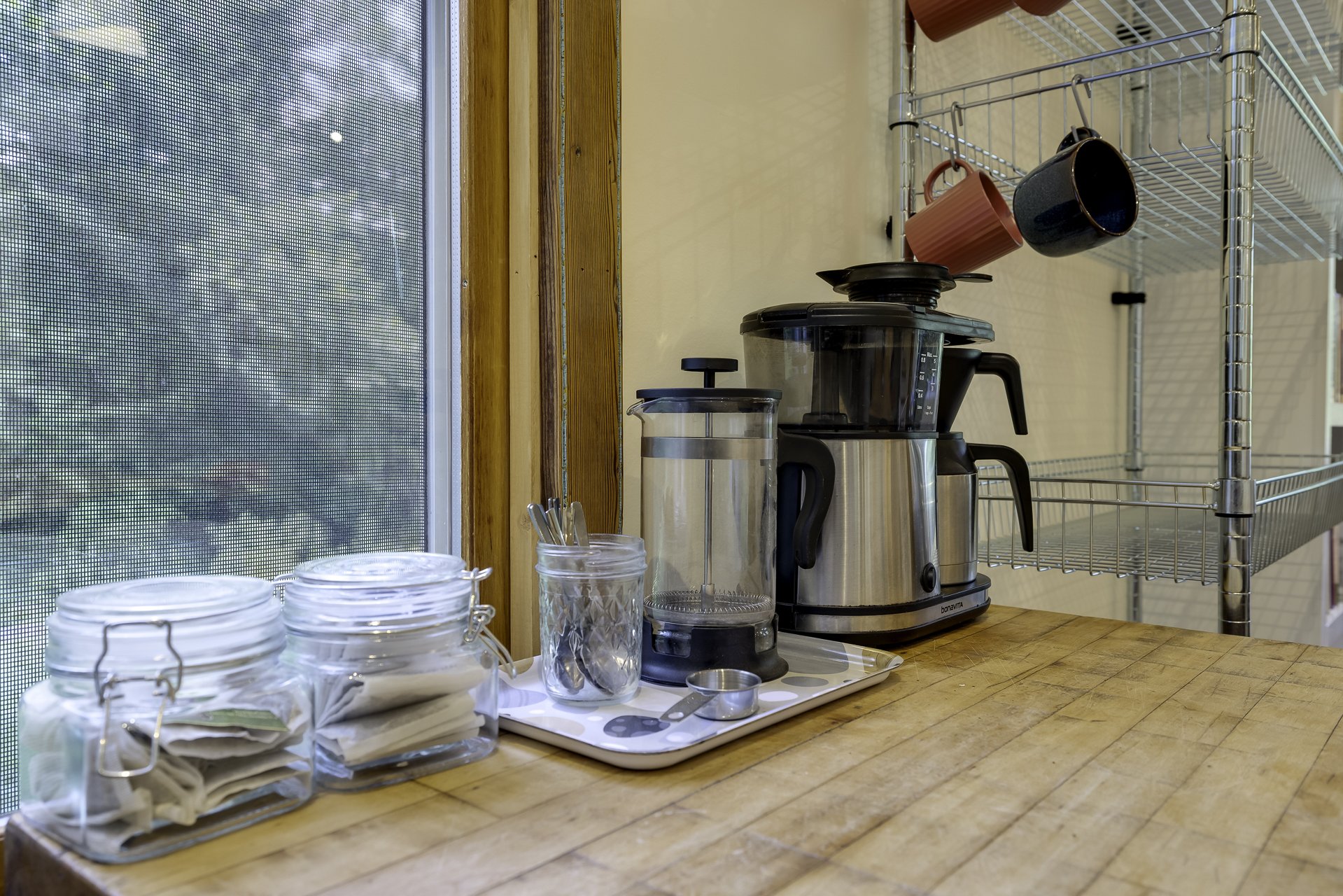 Kitchen countertop with jars, coffee maker, blender, and metal shelf with cups in a cozy kitchen.
