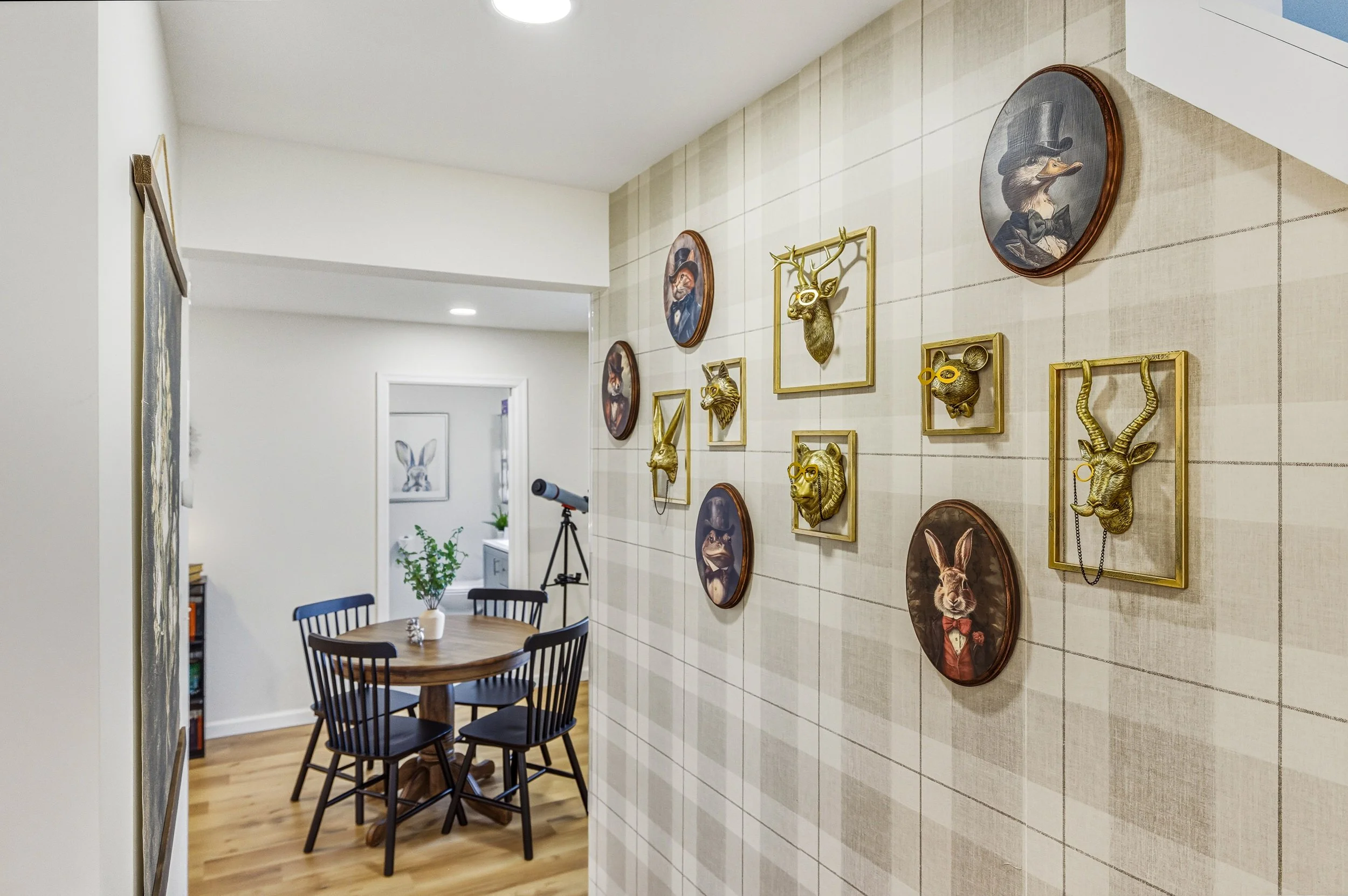 A dining area with a round wooden table and four black chairs. The wall has decorative animal wall art, including framed animal heads and portraits of animals in vintage clothing, such as a fox and a duck, mounted on a tiled wall. There is a potted p