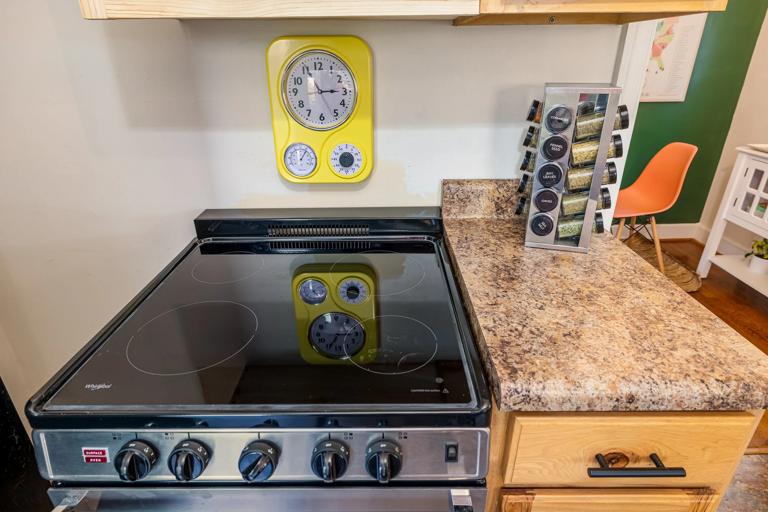 A kitchen stove with four burners and control knobs, part of a granite countertop, with a yellow wall clock and spice rack containing various herbs and spices nearby.