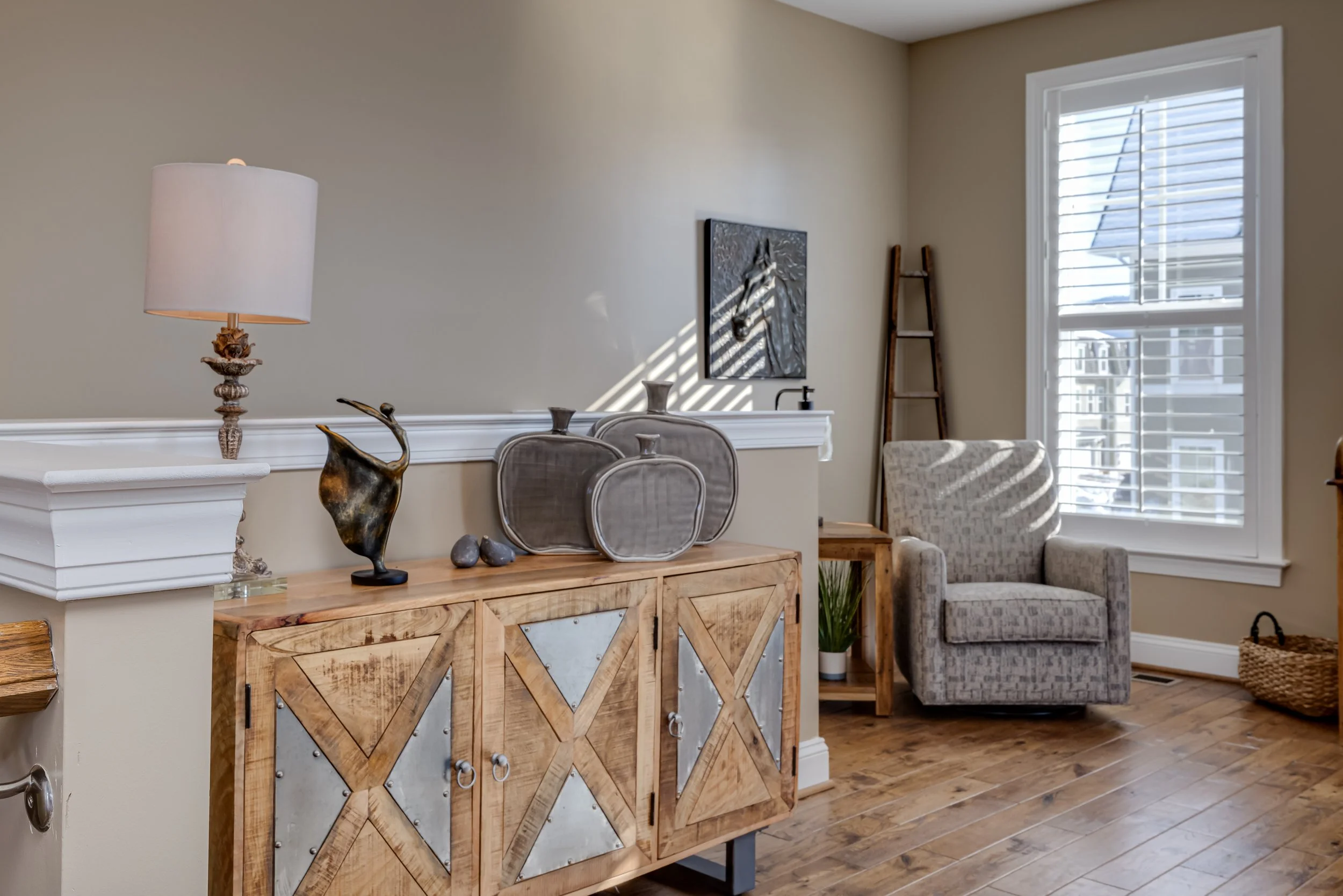 Living room with a wooden sideboard, dragon sculpture, table lamps, gray patterned armchair, window with shutters, ladder, and decorative items.