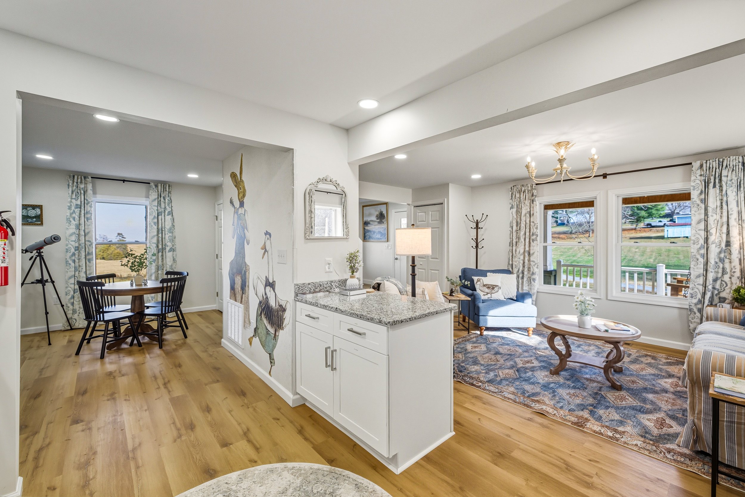 Living room and dining area with large windows, wood flooring, and a chandelier, decorated with artwork and furniture.