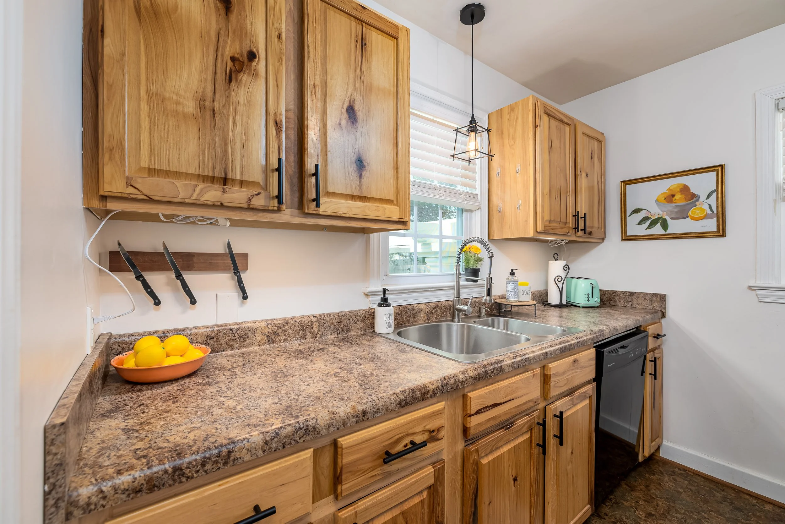 Kitchen with wooden cabinets, a granite countertop, a window with blinds, a double sink, a hanging light fixture, a fruit bowl with lemons, and various small appliances including a soap dispenser, a paper towel holder, and a teal toaster. There is al
