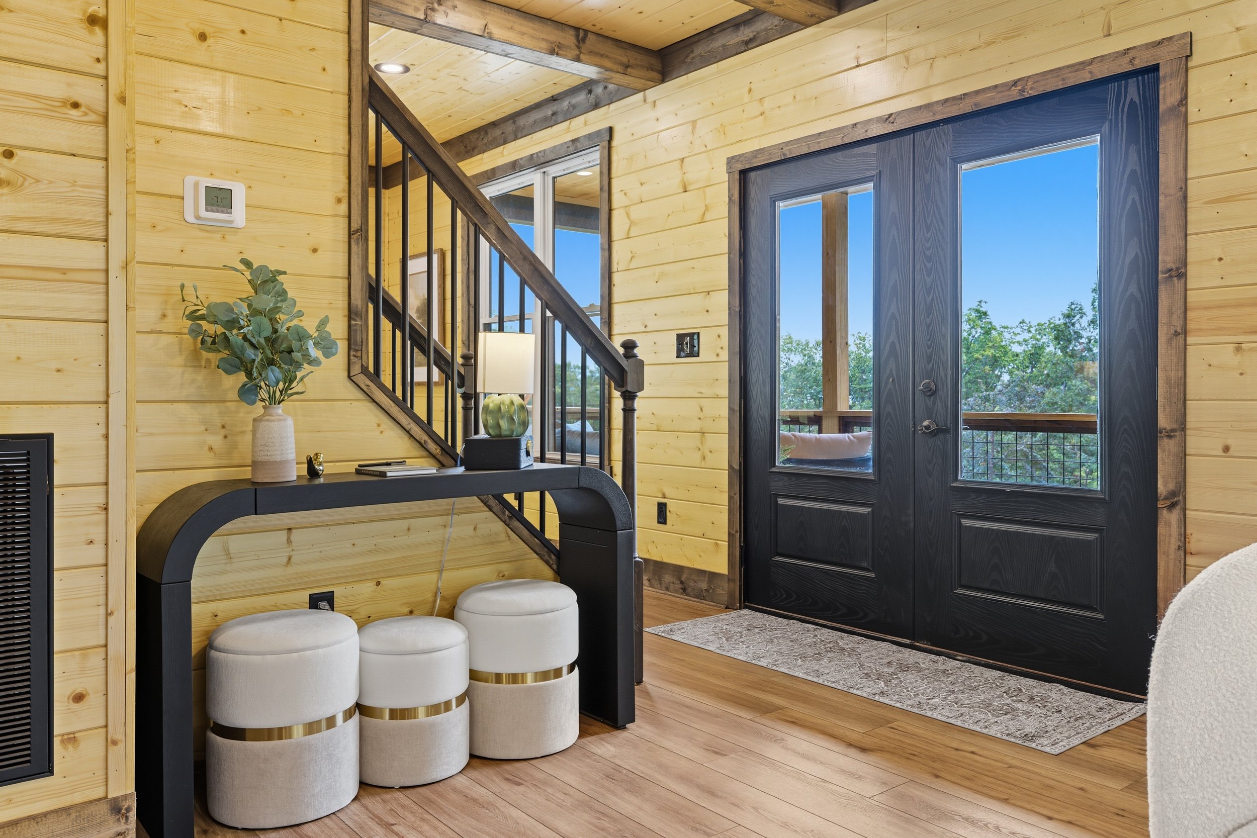 Interior of a wooden cabin with a black door, small window, staircase, a side table with a lamp and decorative items, and three white round ottomans.