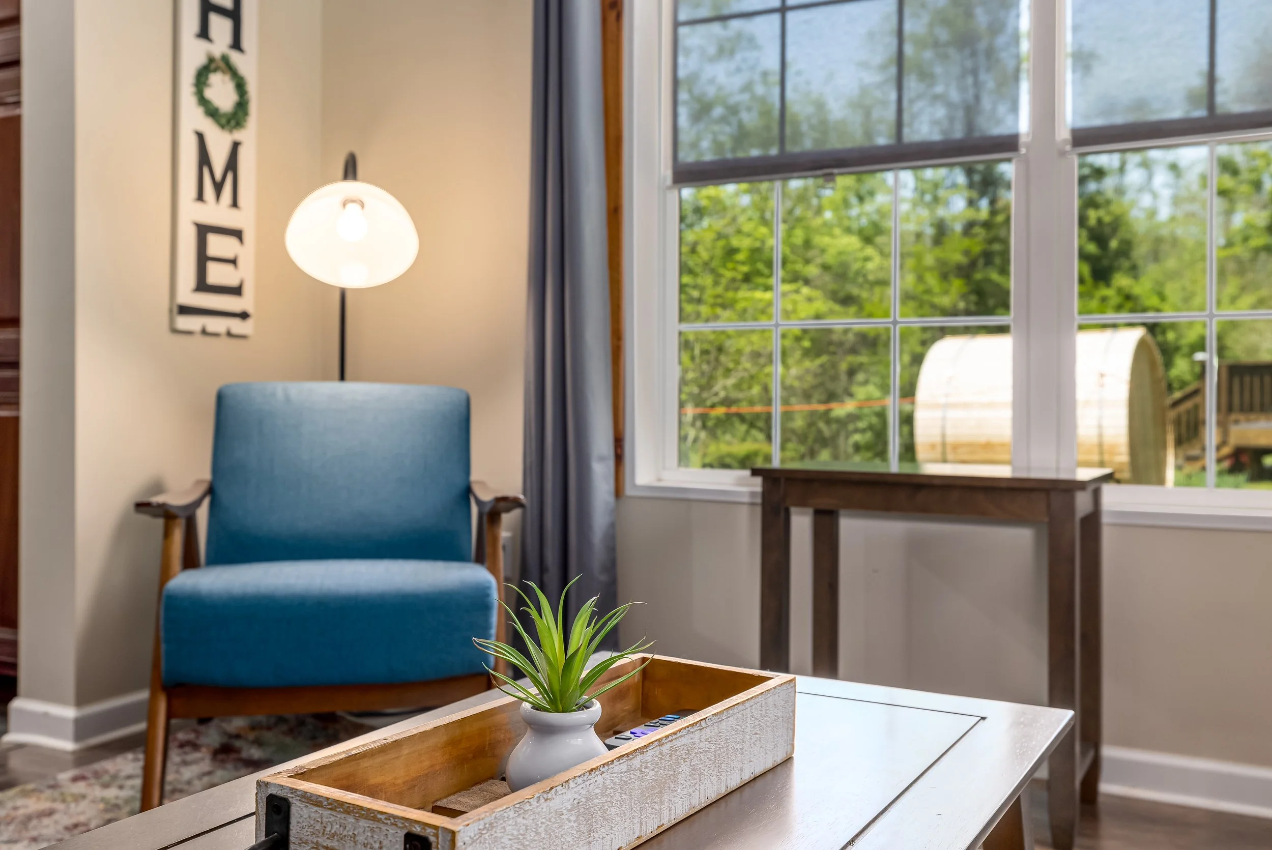 Living room with a blue armchair, a wooden table with a white planter holding a green plant, large windows showing trees outside, a lamp with a round white shade, and wall decor spelling 'HOME' with a green wreath replacing the letter 'O'.