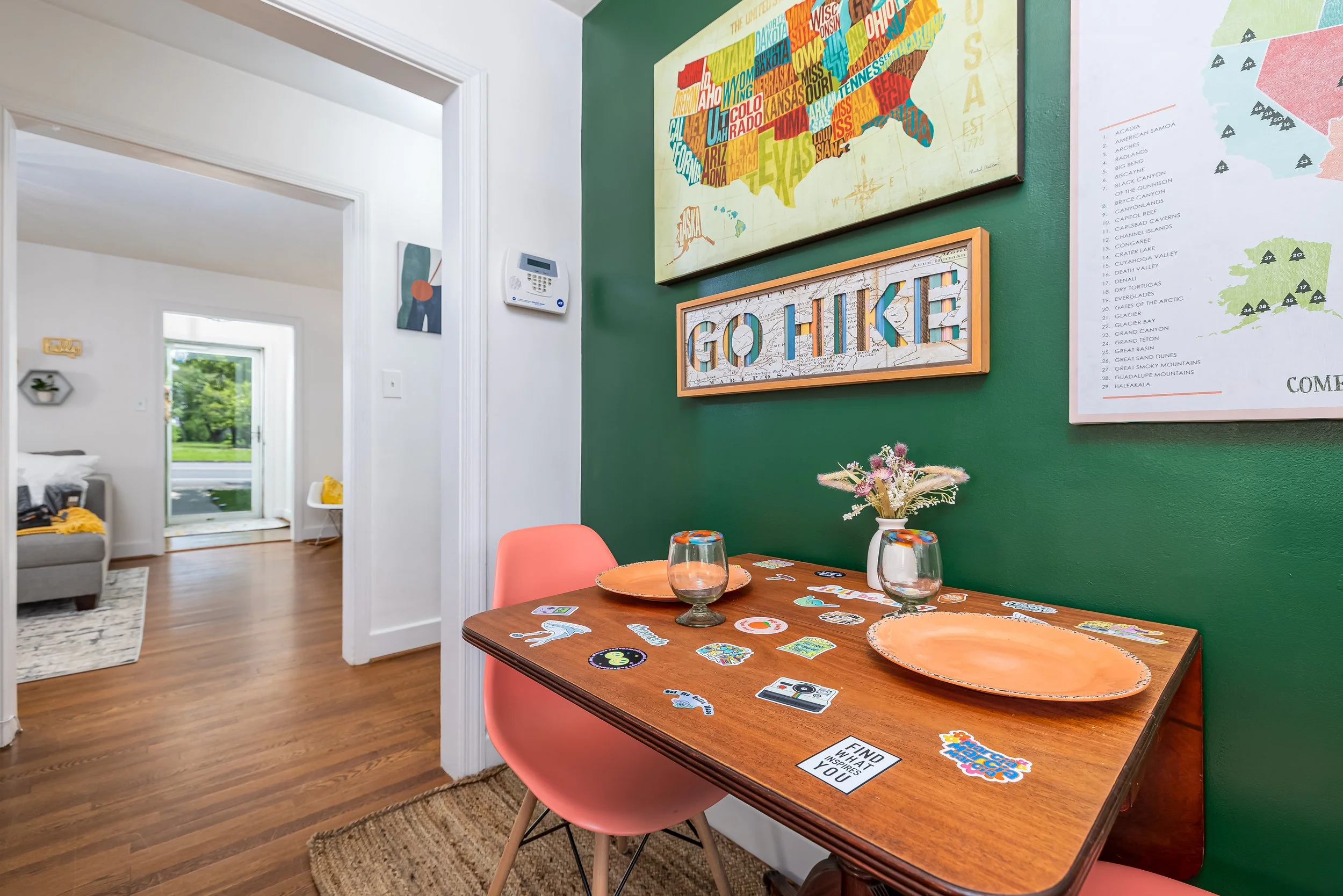 Interior of a home with a green accent wall, decorated with World Map and Alaska Map art and a framed 'Go Chickadees' sign. A wooden dining table with pink chairs is set with two empty glasses and colorful stickers. In the background, there's a white