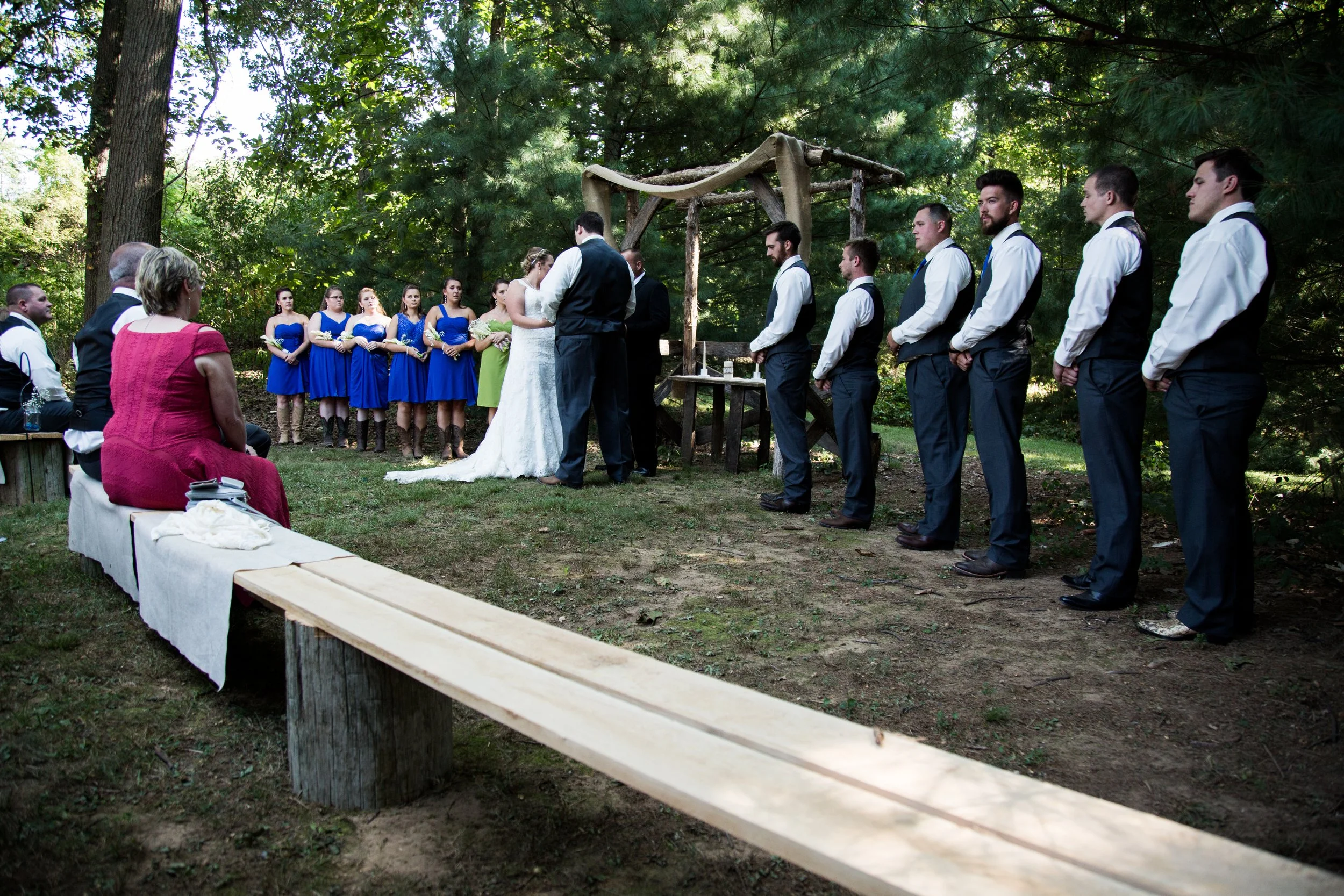 A wedding ceremony taking place outdoors in a wooded area, with the bride and groom standing in front of a wooden arch, surrounded by wedding party and guests.