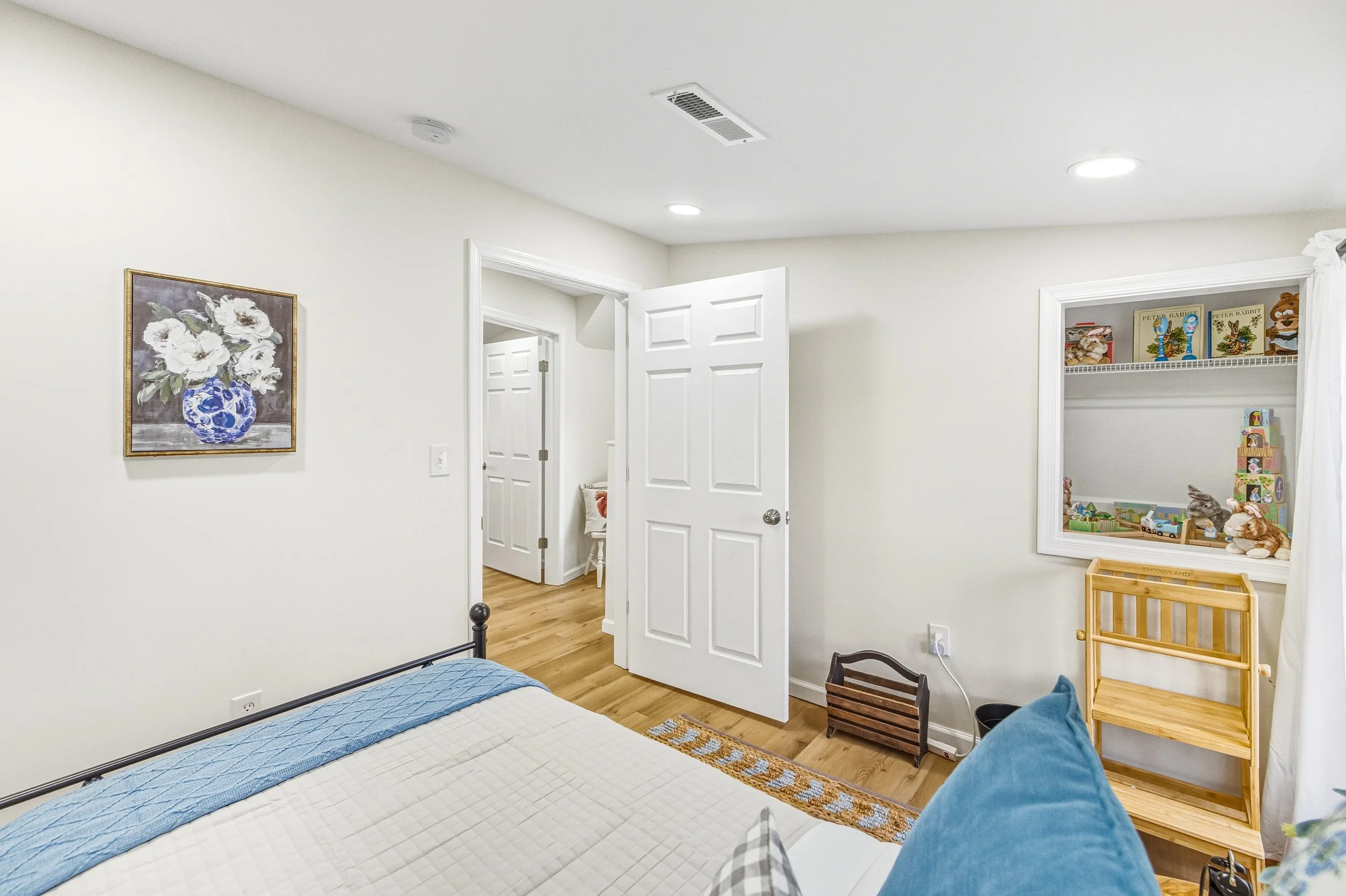 A bedroom with white walls, wooden flooring, and a bed with a blue blanket. There's a painting of white flowers in a blue vase on the wall, and a closet with toys and books inside. The ceiling has recessed lighting.