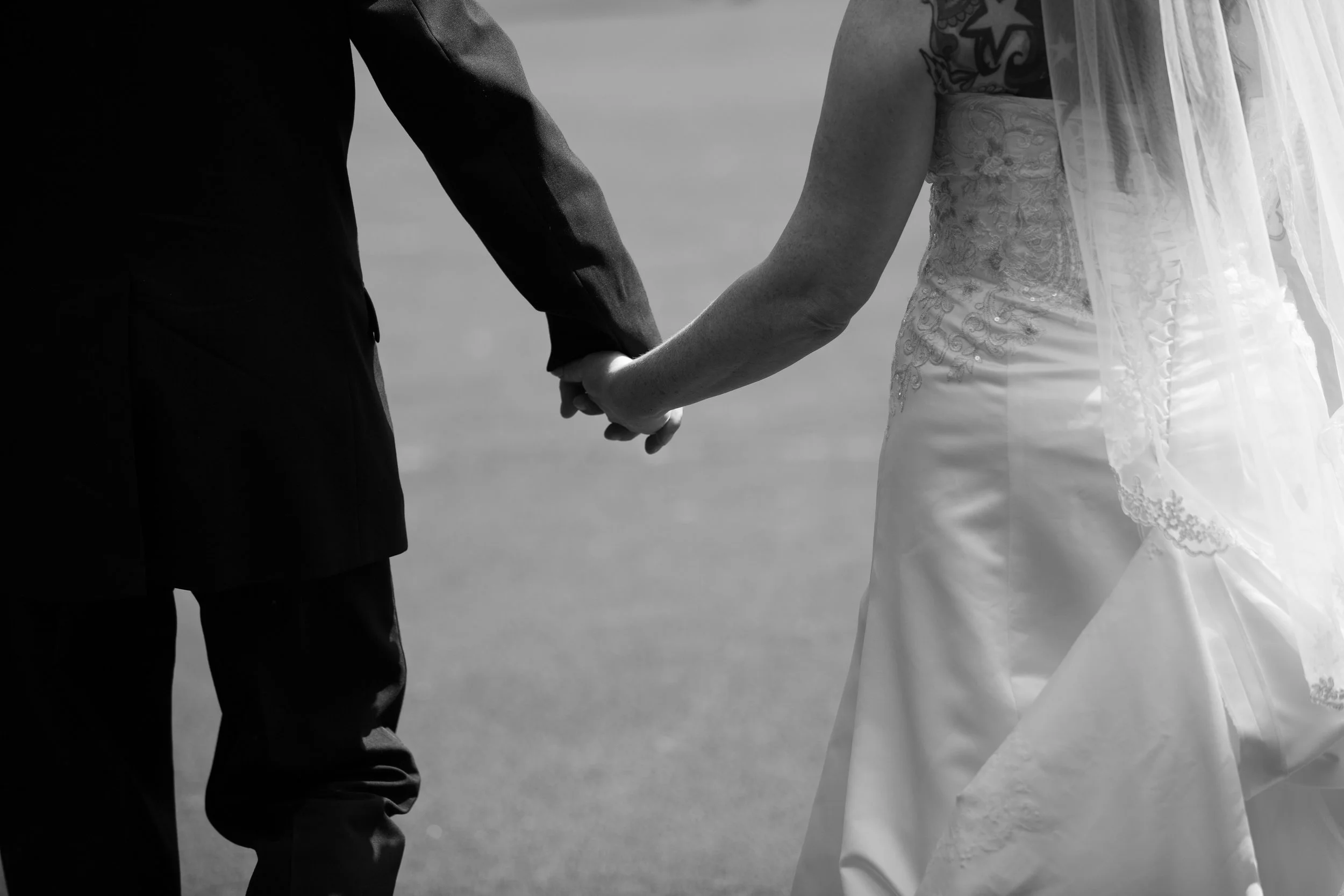 A couple holding hands, dressed in wedding attire, standing outdoors.