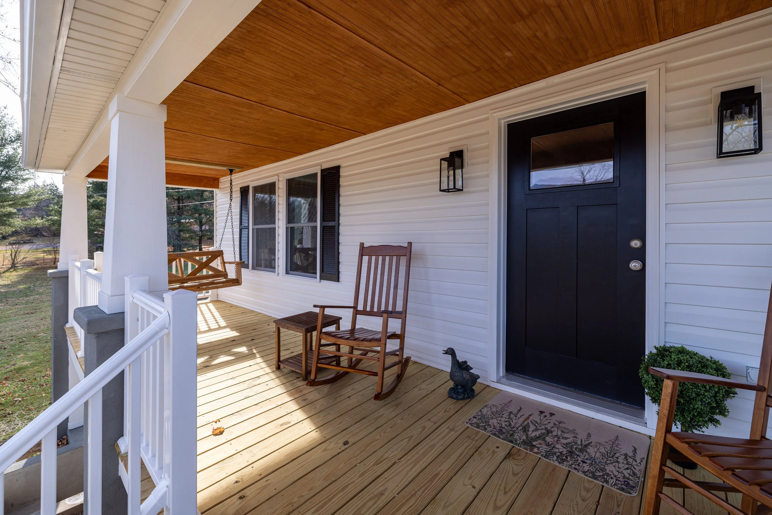 Covered porch with wooden flooring, white siding, black front door, black wall lanterns, and wooden rocking chairs on a sunny day.
