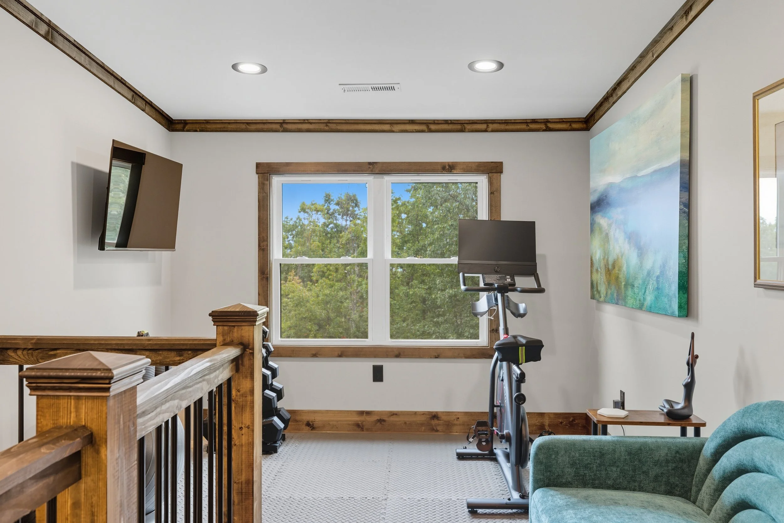 A home gym with a window showing trees outside, a treadmill, a stationary bike, a green armchair, a wall-mounted TV, and a colorful abstract painting.