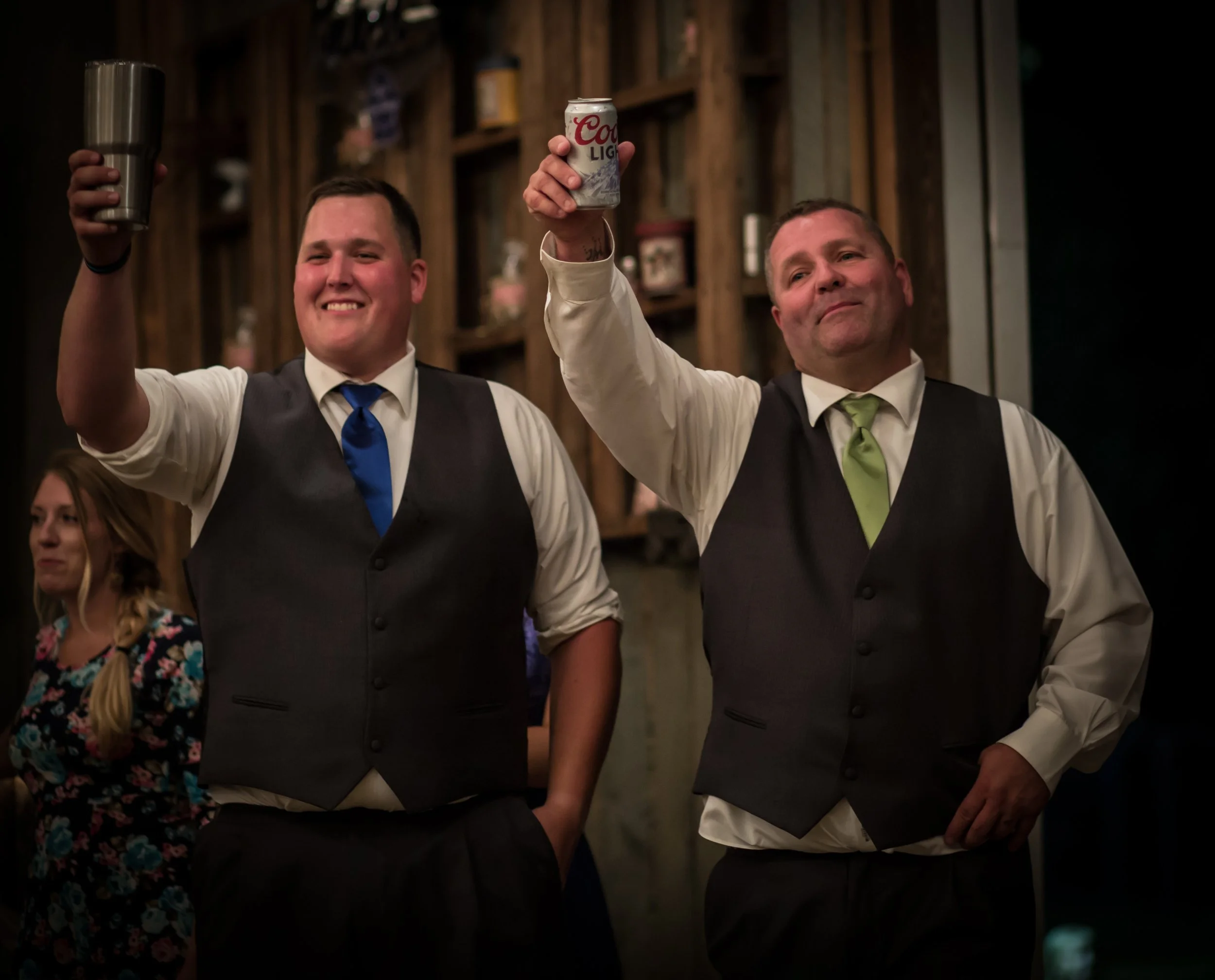 Two men in formal attire raising drinks, smiling, at a celebration or gathering inside a rustic wooden venue.