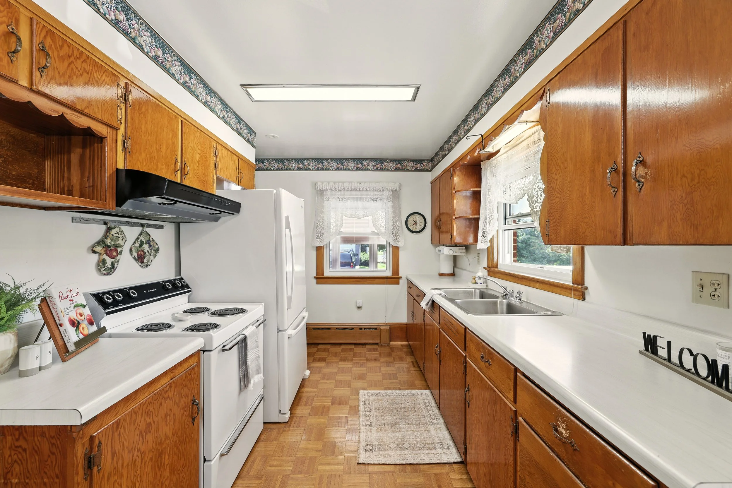 Kitchen with wooden cabinets, white stove, white refrigerator, double sink, and windows with lace curtains.