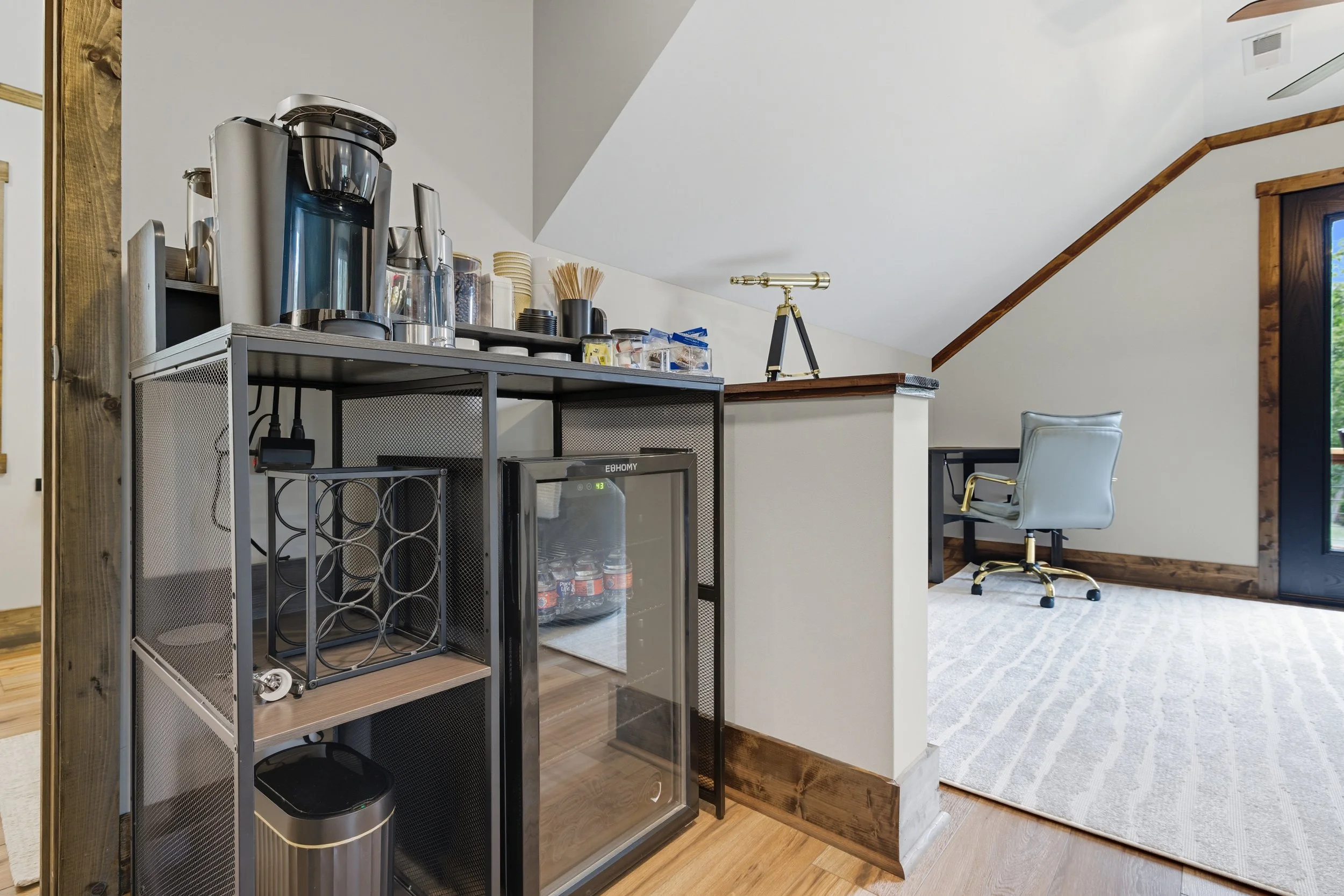 A kitchen area with a coffee station on a metal cart and a small refrigerator, with a white chair and a window with a wooden frame in the background.