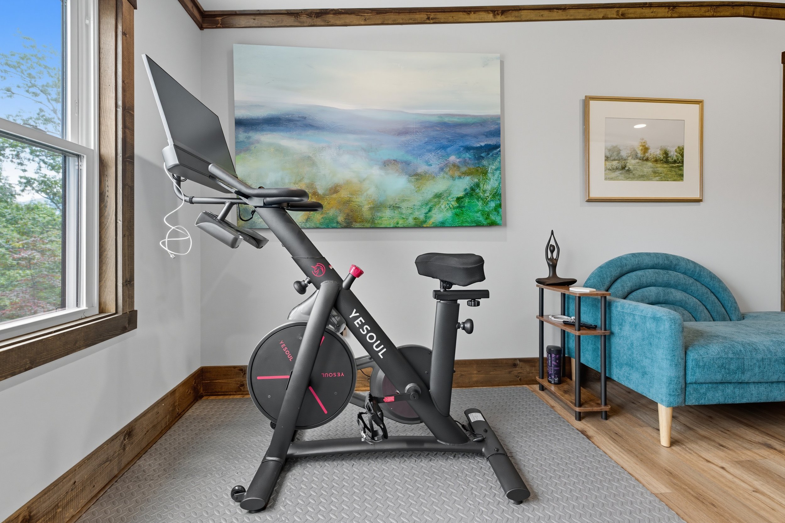A stationary exercise bike is placed in a cozy room with wood accents and artwork on the walls. There are two paintings, one larger abstract landscape and a smaller framed landscape scene, hanging on the white walls. A blue upholstered chair and smal