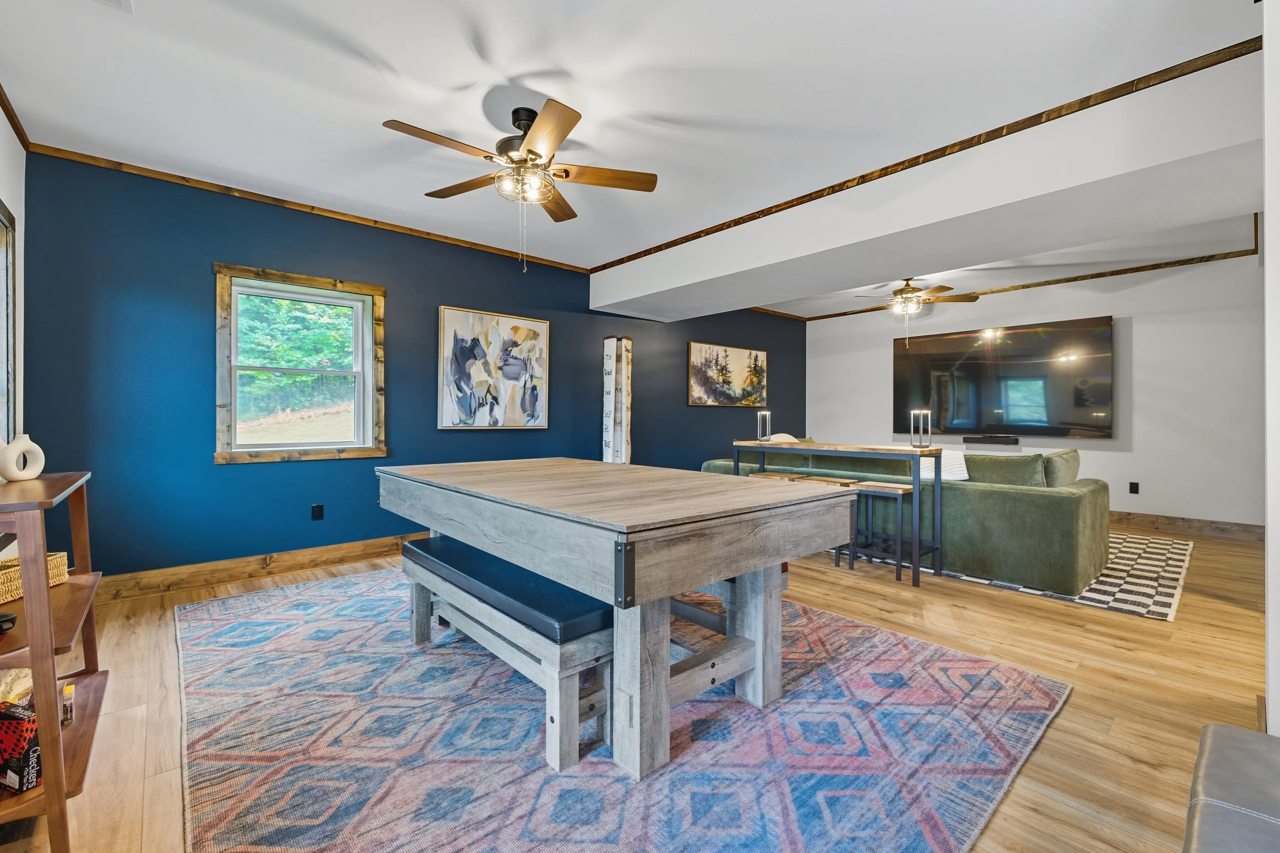 Living room with blue accent wall, wooden trim, two ceiling fans, artwork, a TV on the white wall, and a wooden dining table with a bench, rug, and sofa.