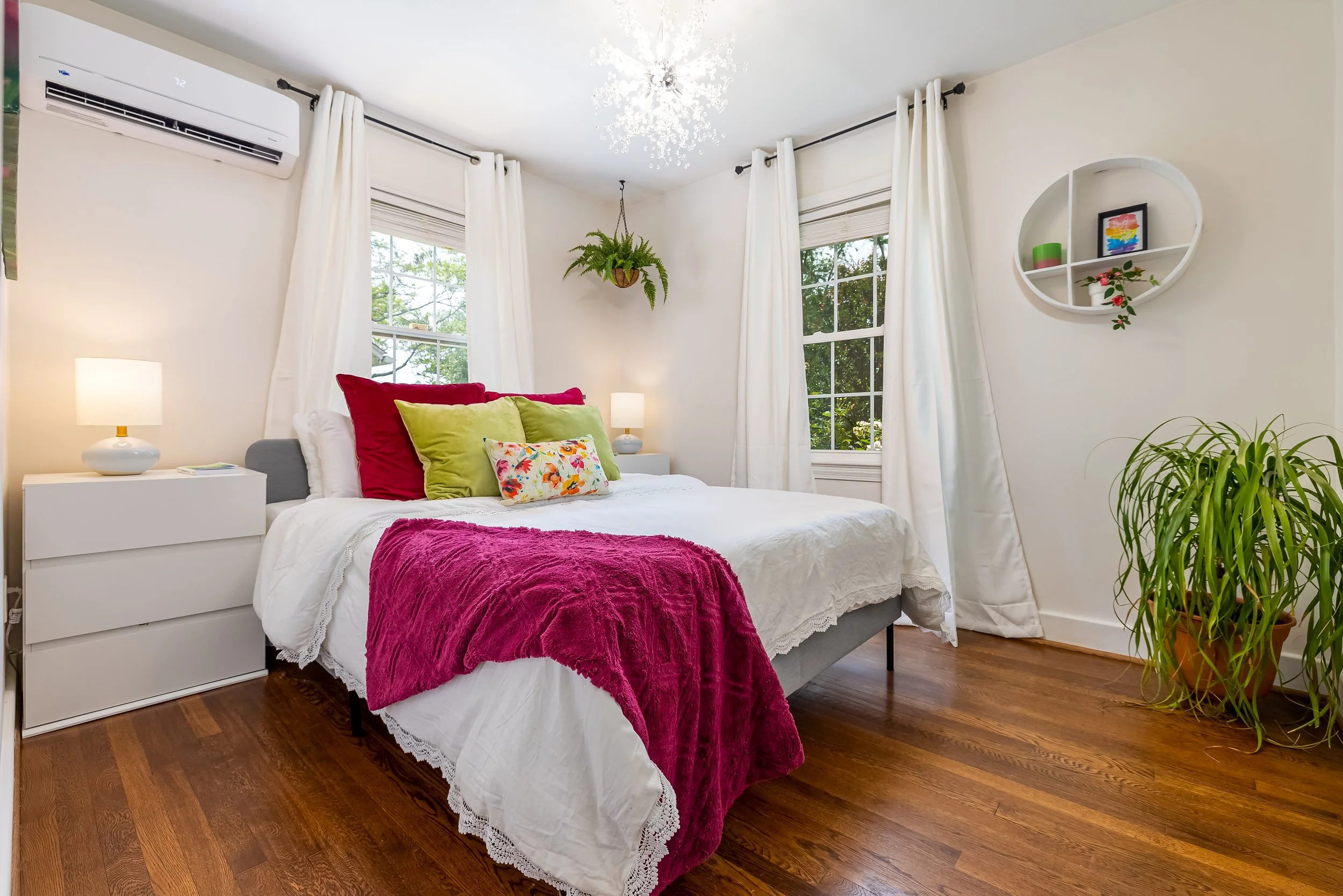 A cozy bedroom with white walls, white curtains, a bed with white linens, colorful pillows, and a pink throw blanket. There are two windows with trees outside, a nightstand with a lamp, a hanging plant, a decorative wall shelf, and a hardwood floor.