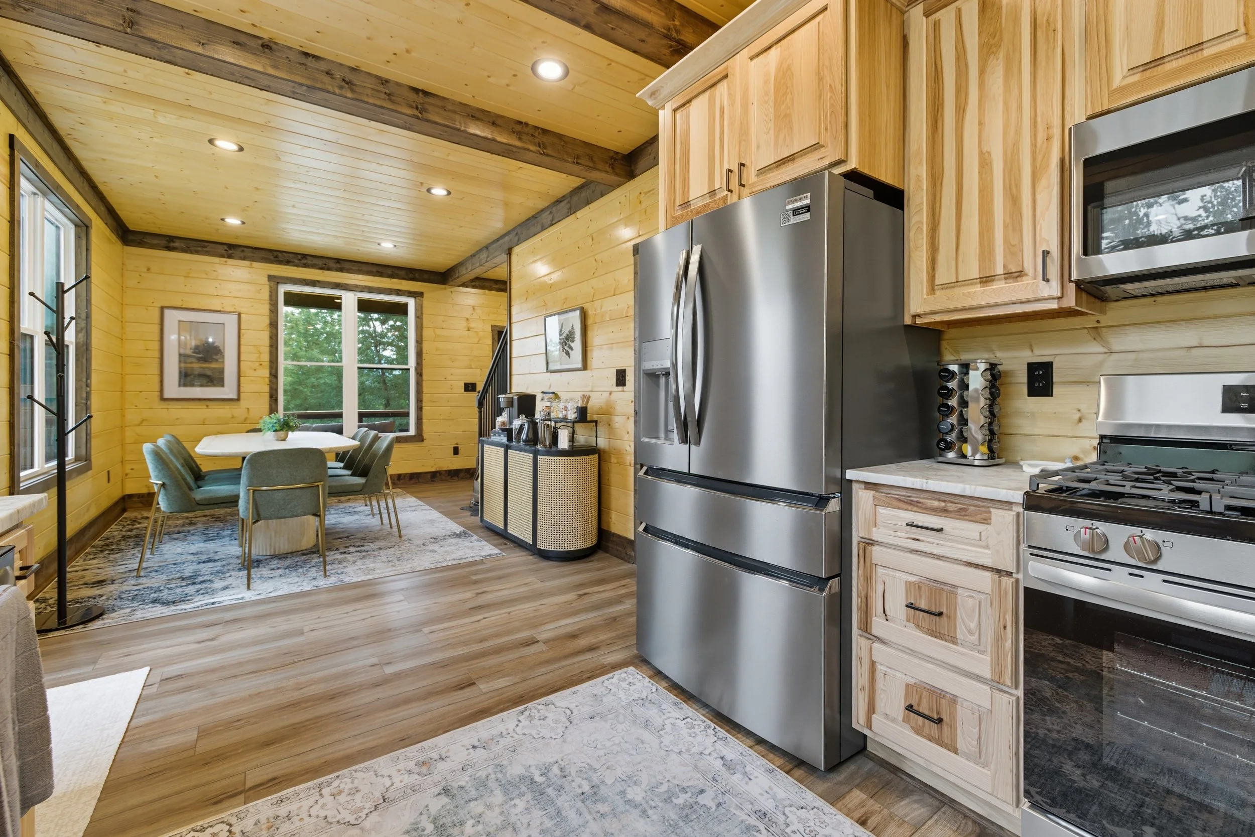 Interior view of a wooden kitchen and dining area with a stainless steel refrigerator, microwave, gas stove, and a dining table with chairs. The room features wood-paneled walls and ceiling, large windows, and framed artwork.