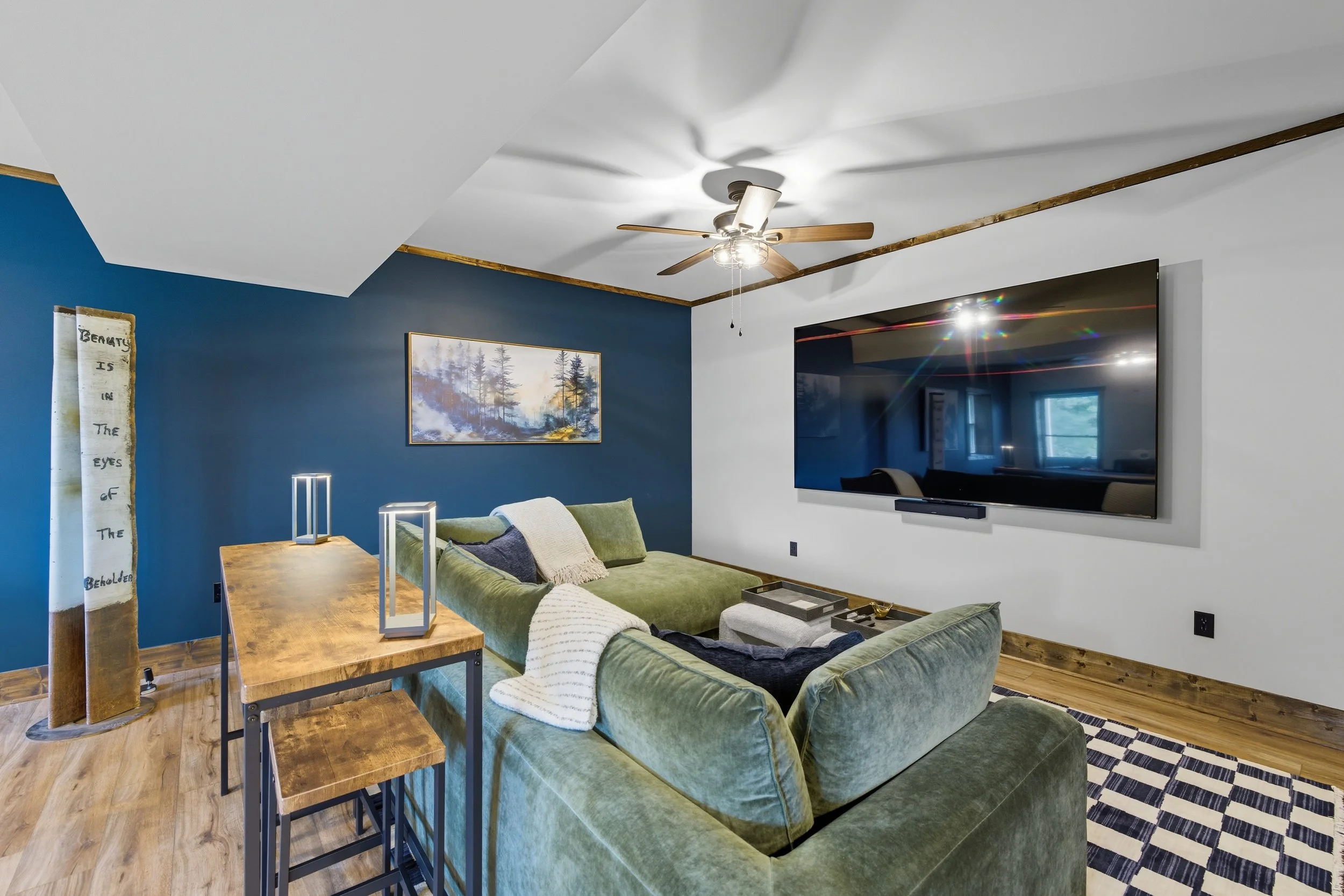 A living room with a green velvet sofa, a wall-mounted television, a wooden dining table, and blue accent walls, decorated with landscape artwork and modern fixtures.