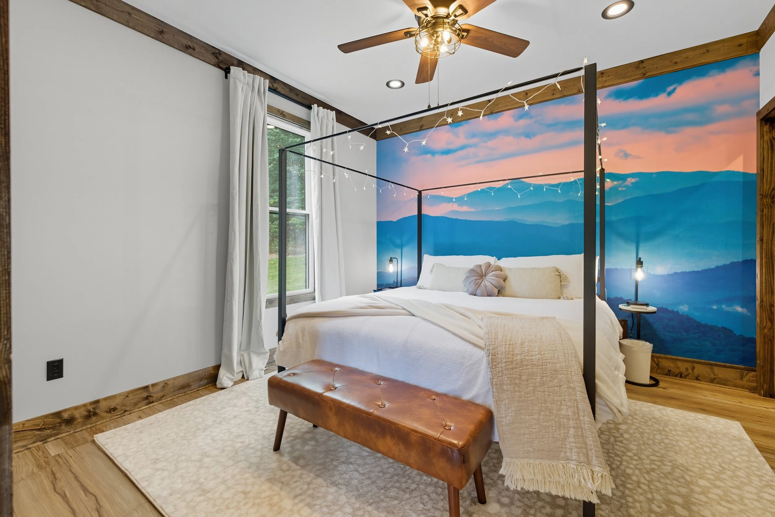 A cozy bedroom with a bed, a brown leather ottoman at the foot, and a mountain mural behind the bed. The room has wooden trim, a ceiling fan, and string lights.