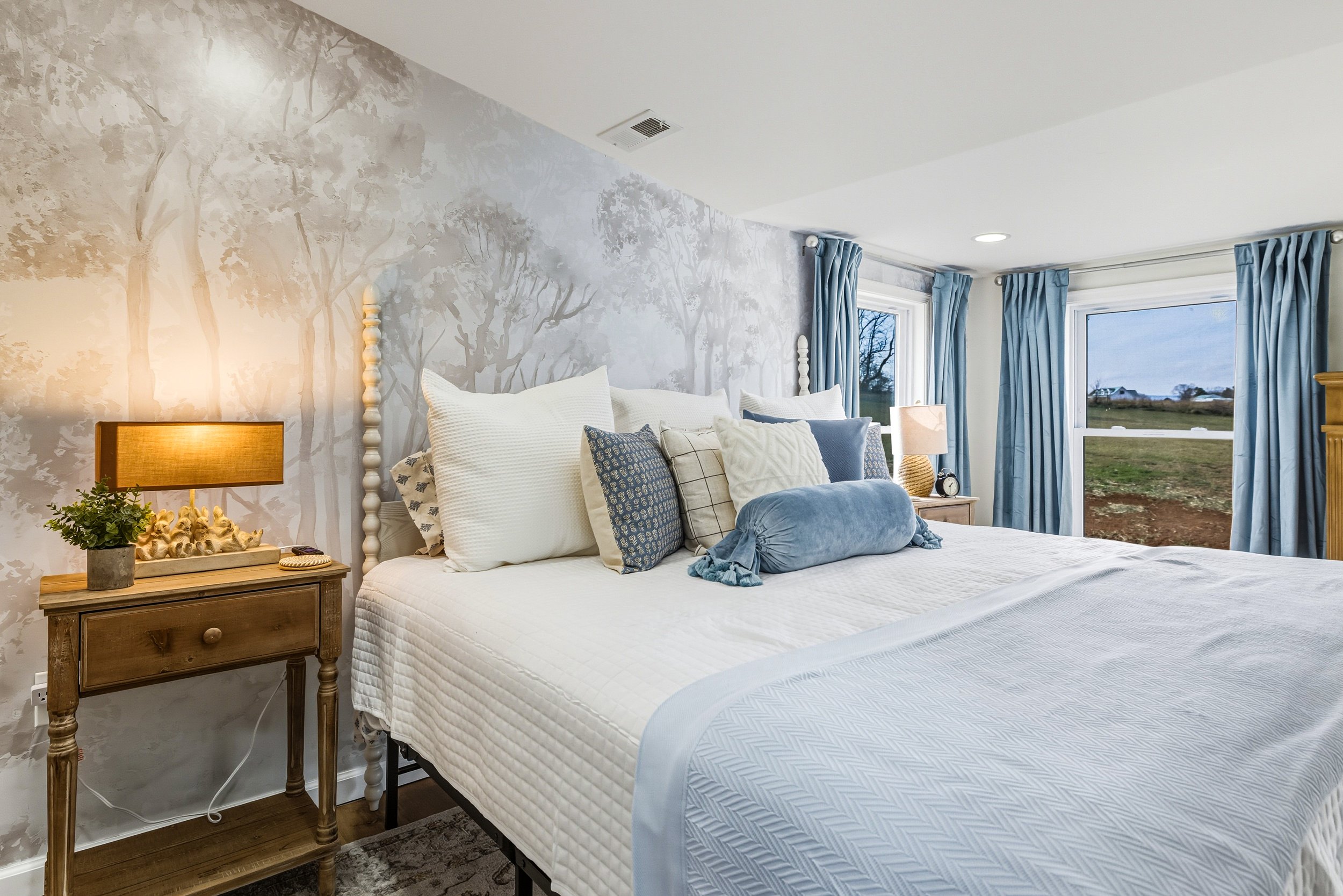 A cozy bedroom with a white bed, multiple pillows, a wooden nightstand with a plant and lamp, and large windows with blue curtains showing an outdoor landscape.