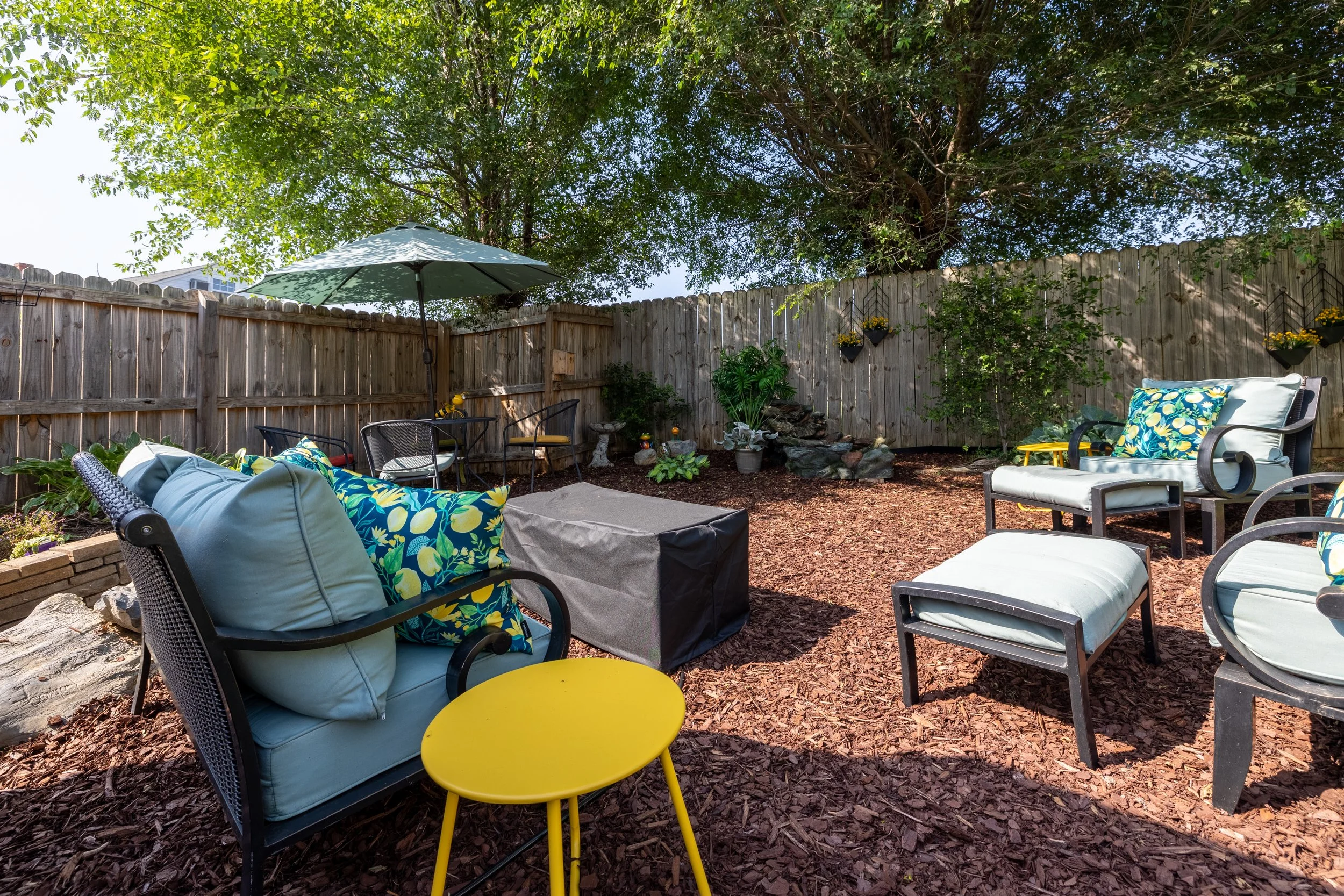 A cozy backyard patio with various seating options, including cushioned chairs with tropical print pillows, a lounge chair, and a small yellow table. There is an umbrella providing shade, surrounded by a wooden fence with hanging plant baskets, lush 