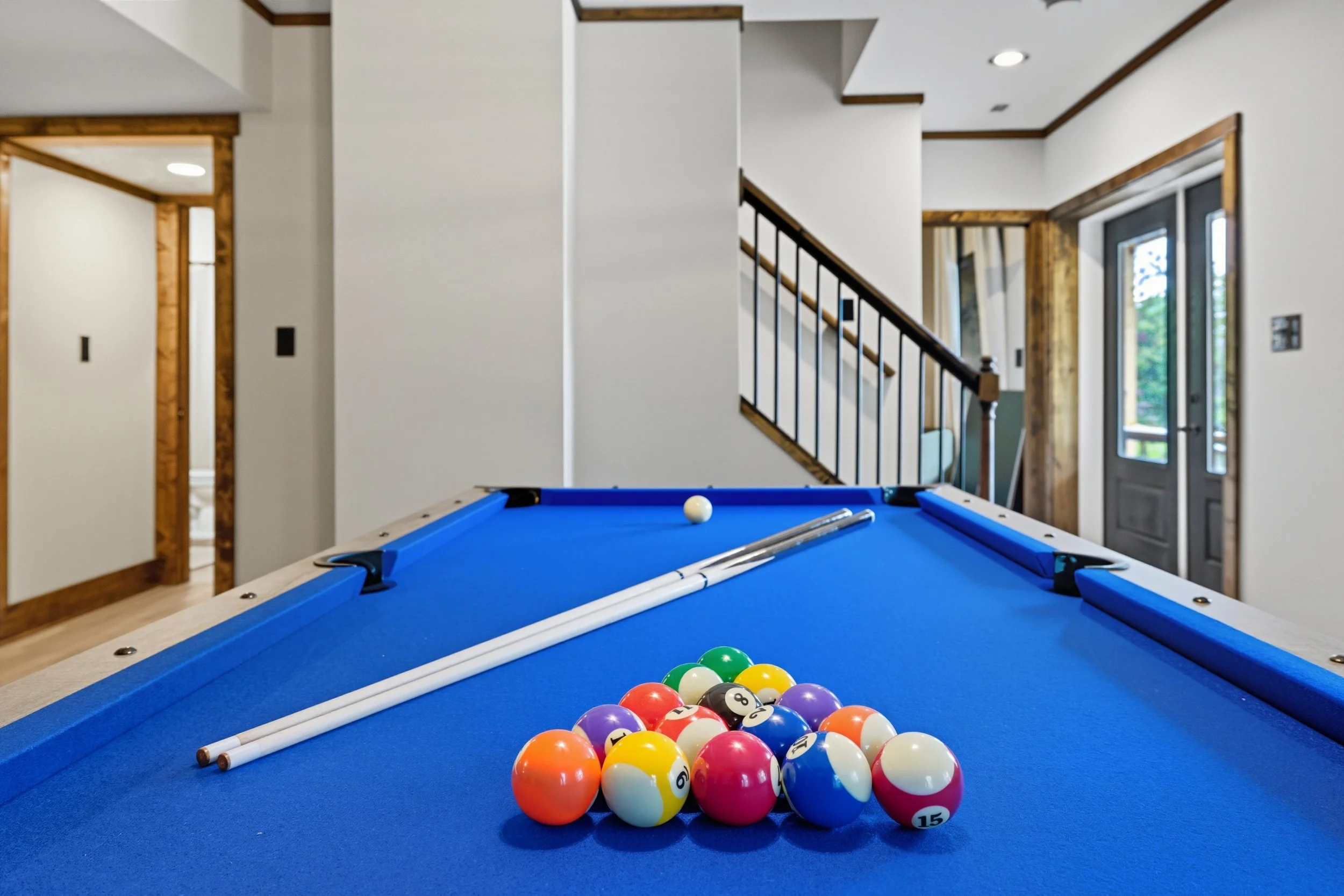 A blue pool table with a rack of pool balls and two pool cues resting on it in a well-lit room with white walls, wooden accents, and large windows