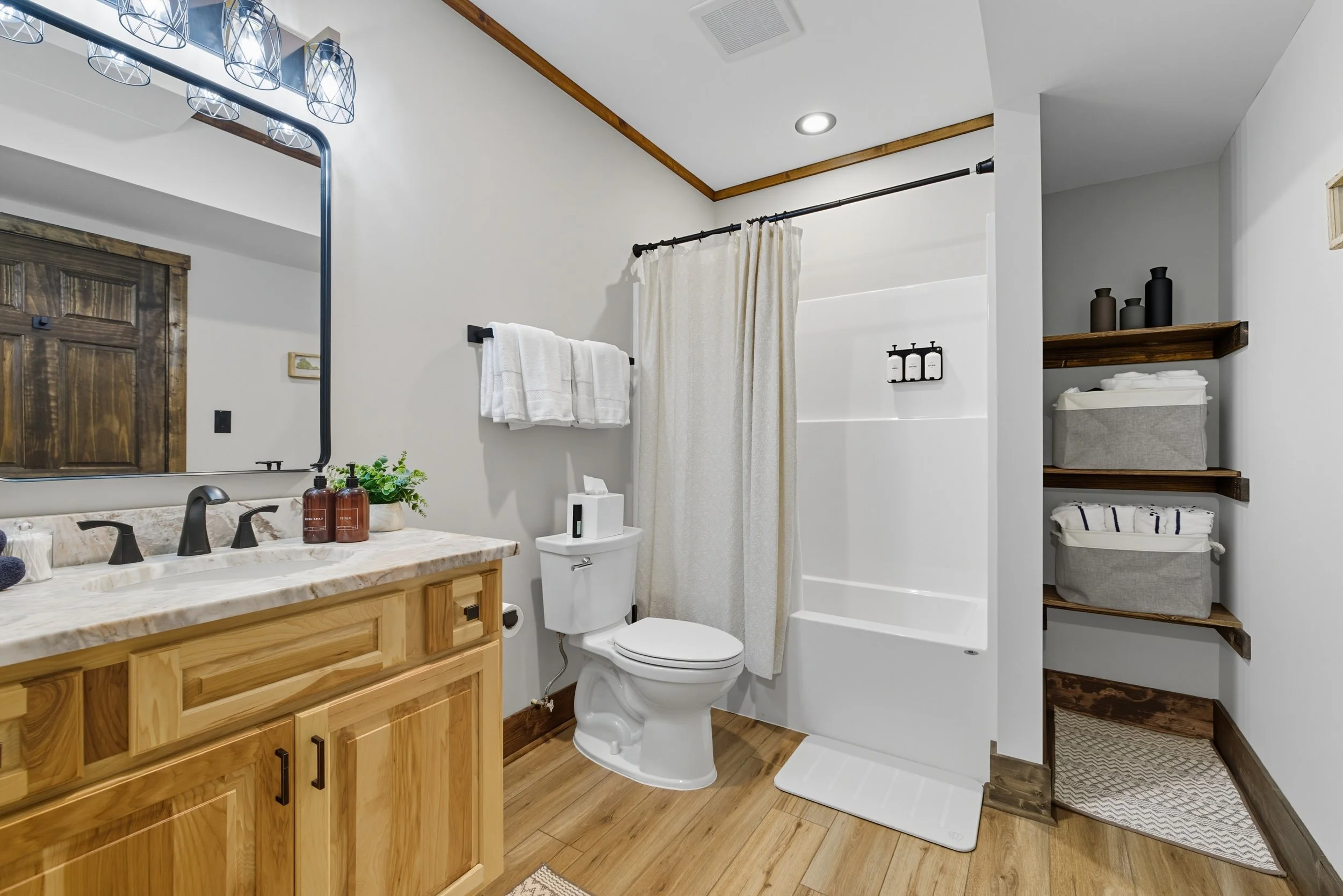 Modern bathroom with wooden vanity, black fixtures, white countertop, and large mirror. Contains towel rack, toilet, shower with curtain, and open shelving with baskets and decor.