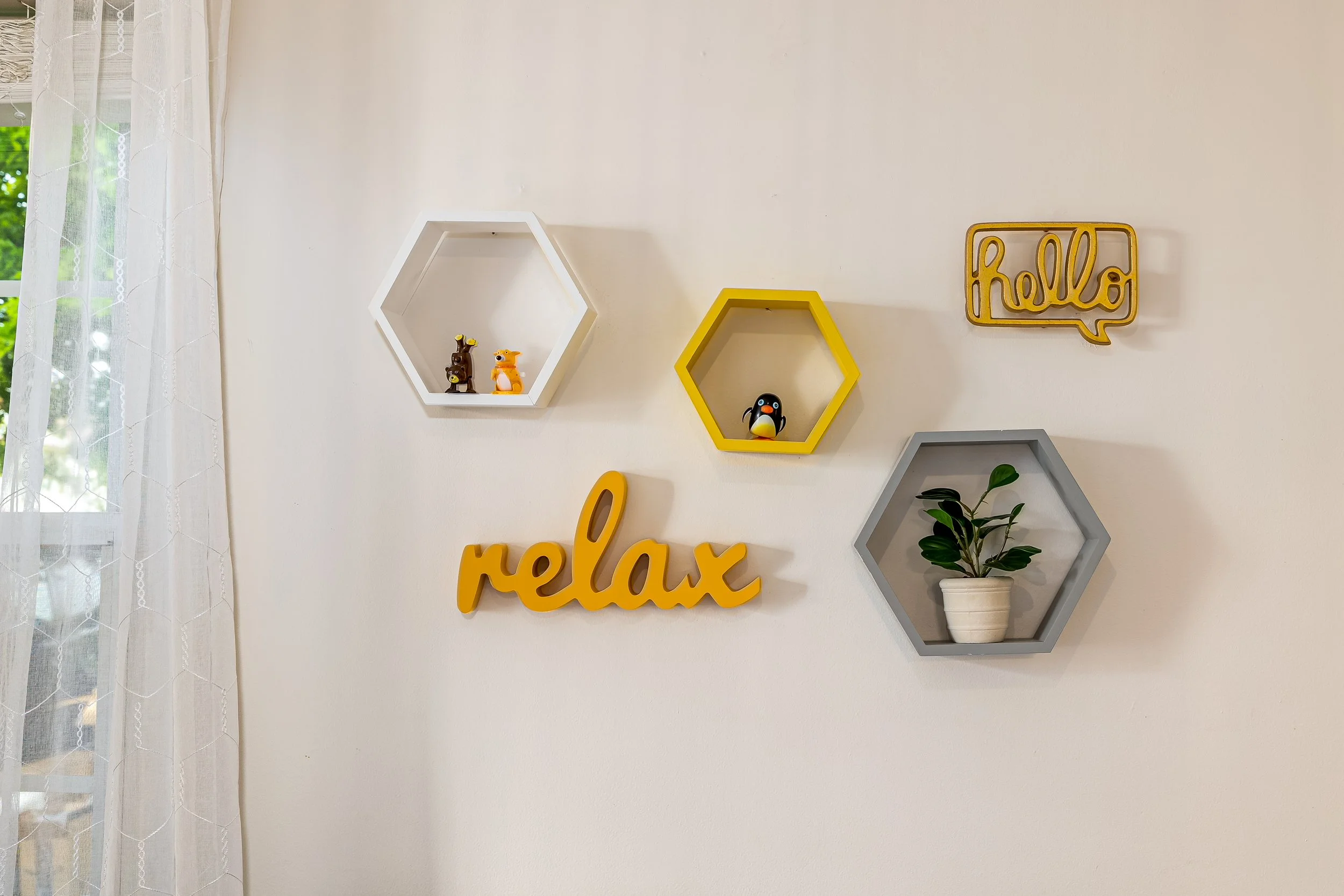 Decorative wall with shelves, a 'relax' sign, a plant, and a 'hello' sign on a light-colored wall next to sheer curtains and a window showing green trees outside.
