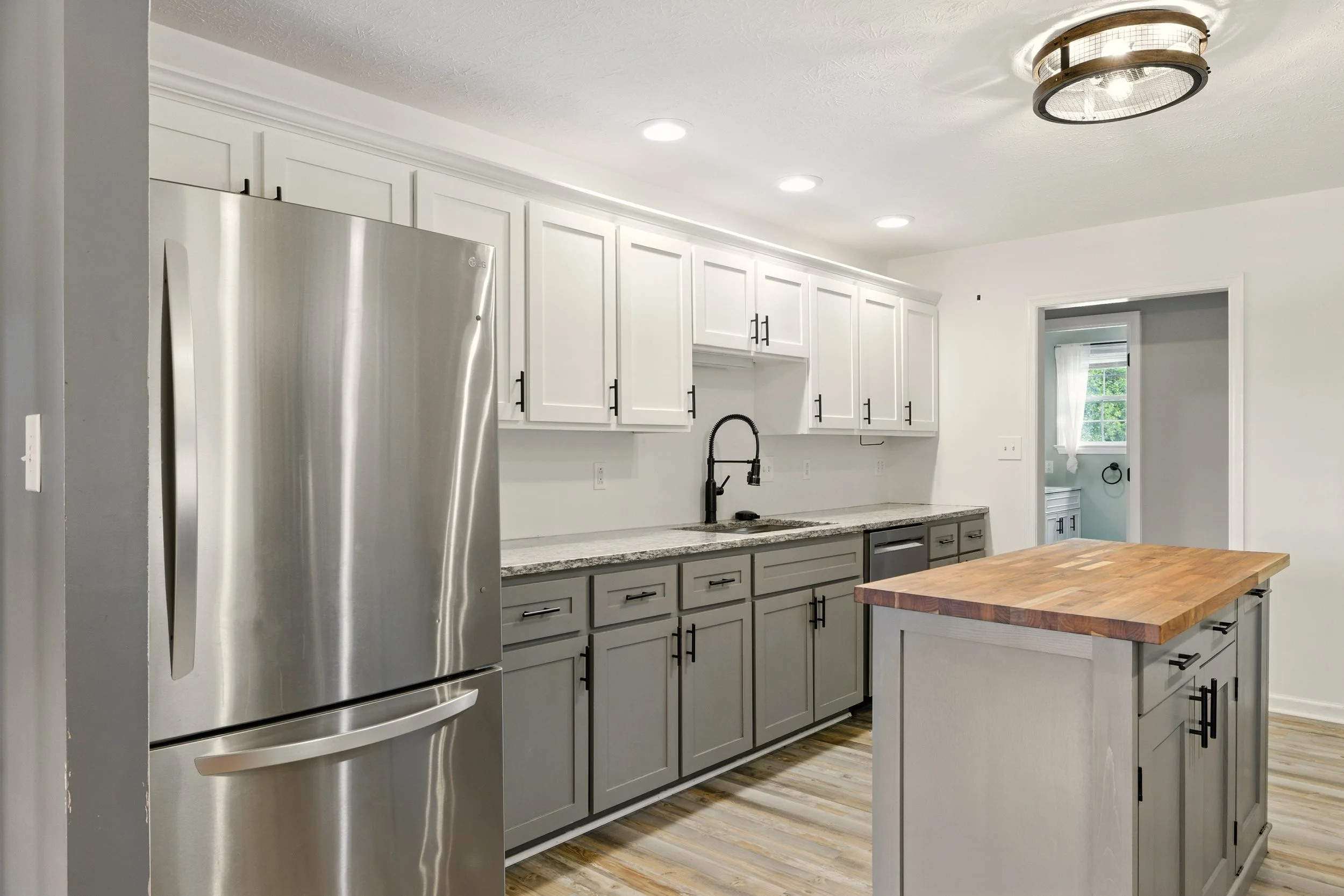 Modern kitchen featuring white upper cabinets, gray lower cabinets, a wooden kitchen island, and stainless steel appliances including a refrigerator. There is a black kitchen faucet and a window with white curtains in the background.