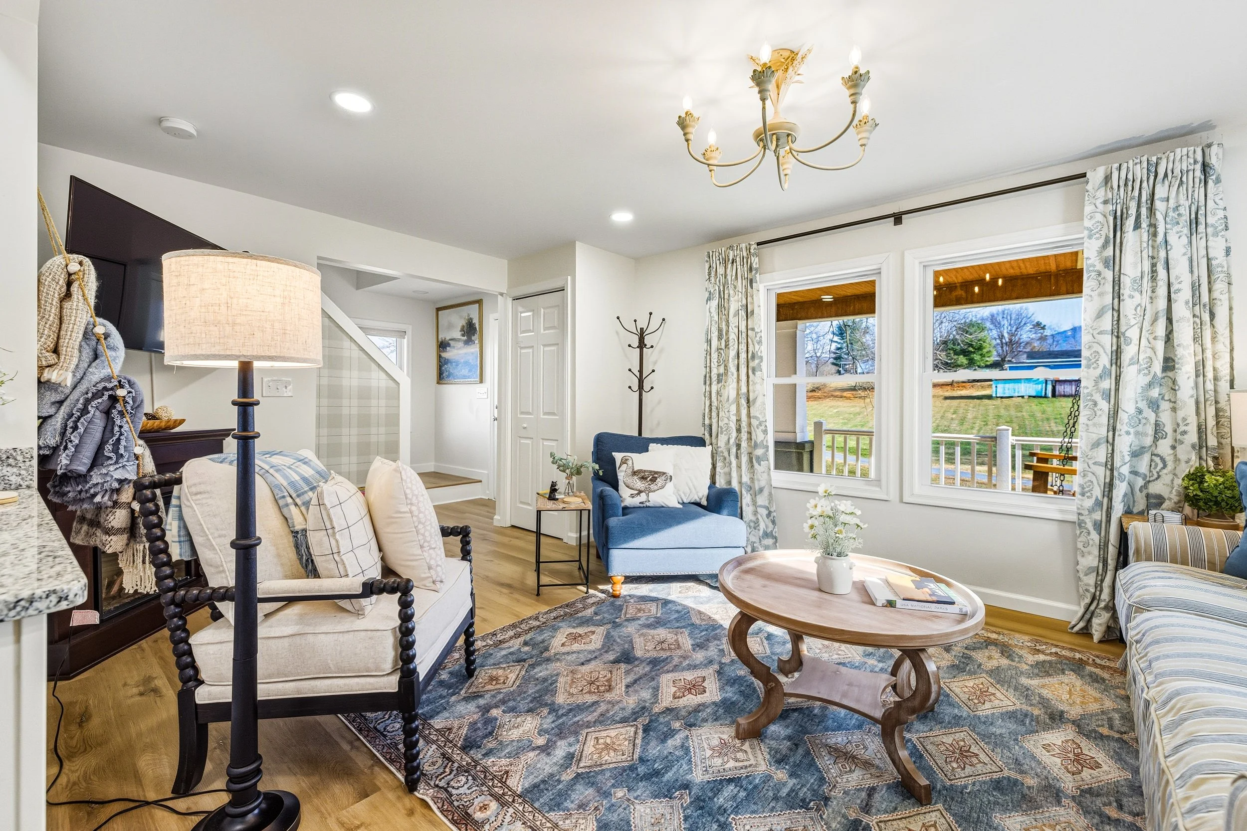 Living room with white walls, large windows with patterned curtains, and hardwood floors. There is a blue armchair with a decorative pillow, a wooden coffee table with a vase of white flowers, and a patterned area rug. A chandelier is hanging from th