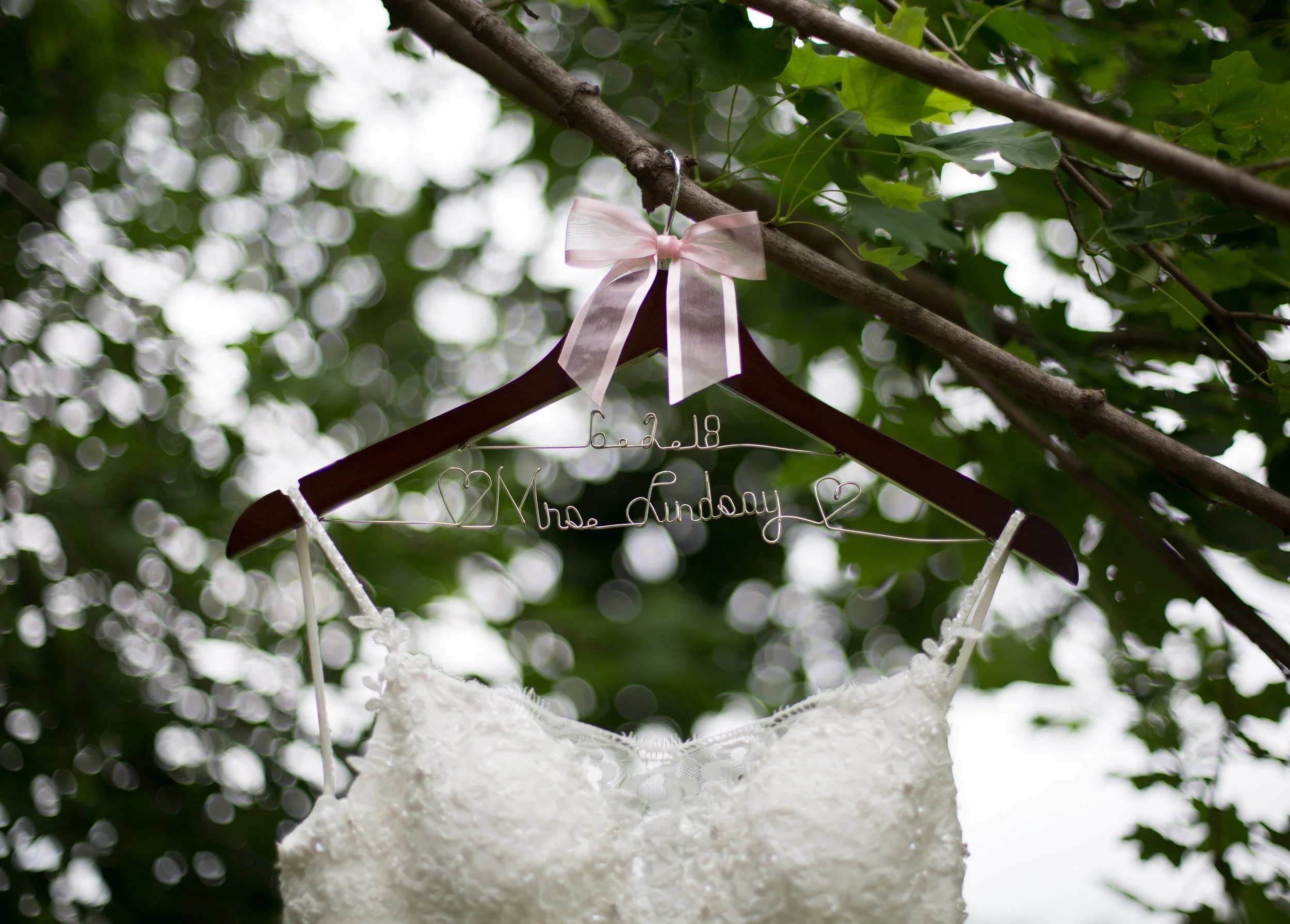 A white dress hanging from a hanger with a pink ribbon bow, suspended from a tree branch with green leaves, with a blurred bokeh background.