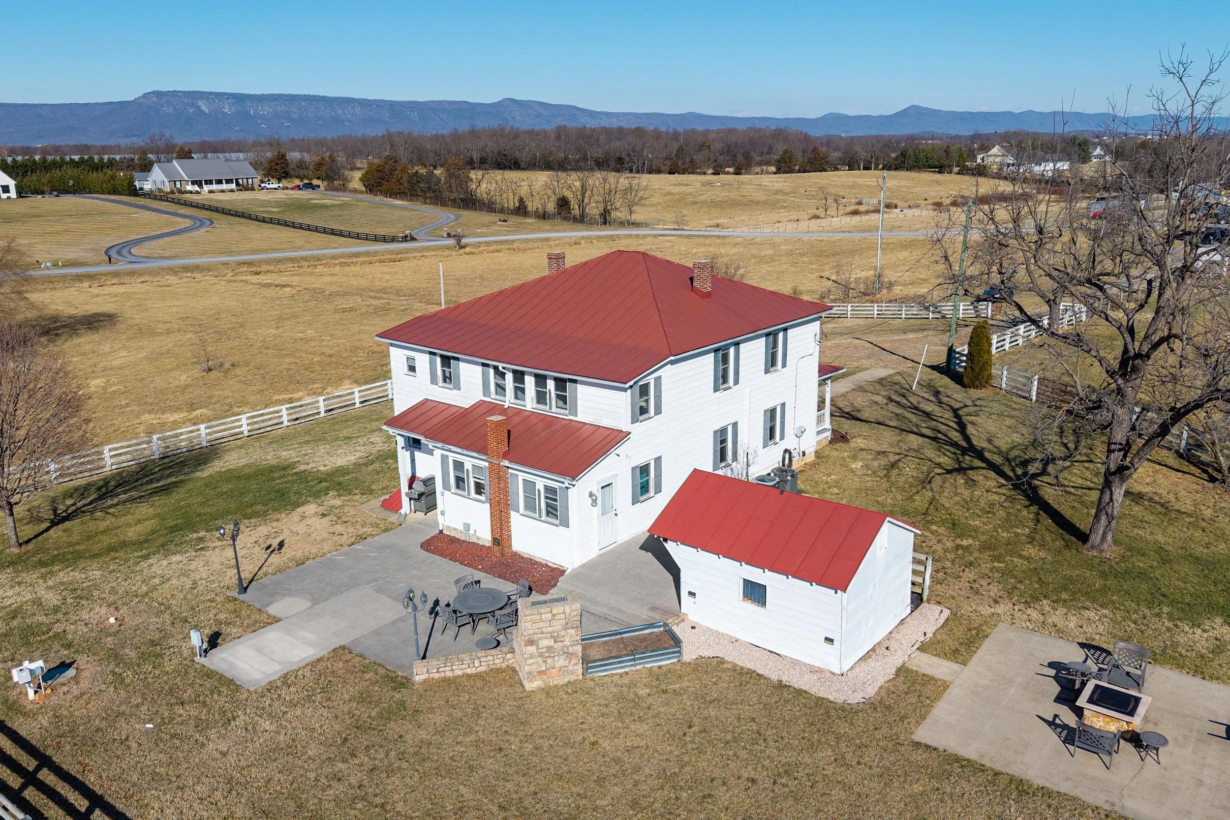 A white two-story house with a red metal roof, a fenced backyard, and a concrete patio with outdoor furniture. The yard has a small garden and a fire pit with seating. There are leafless trees and a large open field with winding roads and distant mou