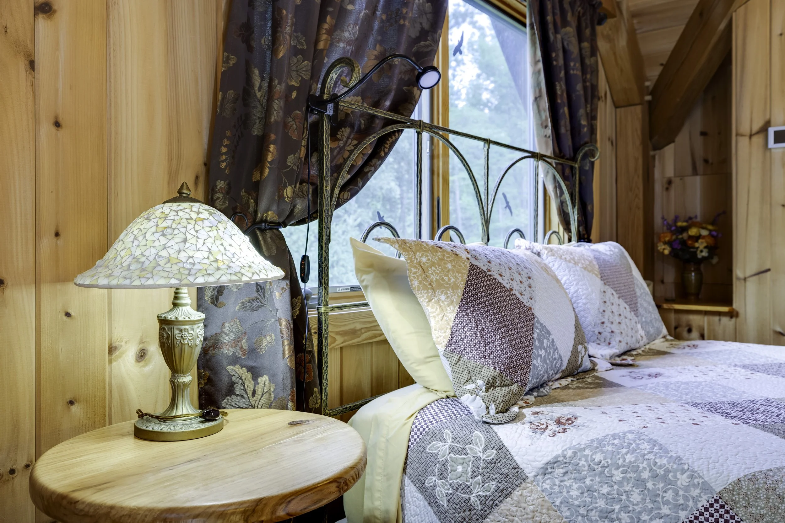 A cozy bedroom with wooden walls and a window showing greenery outside. There is a brass bed with pillows and a patchwork quilt. A round wooden nightstand holds a decorative table lamp with a mosaic glass shade and a small black device. Dark floral curtains frame the window.