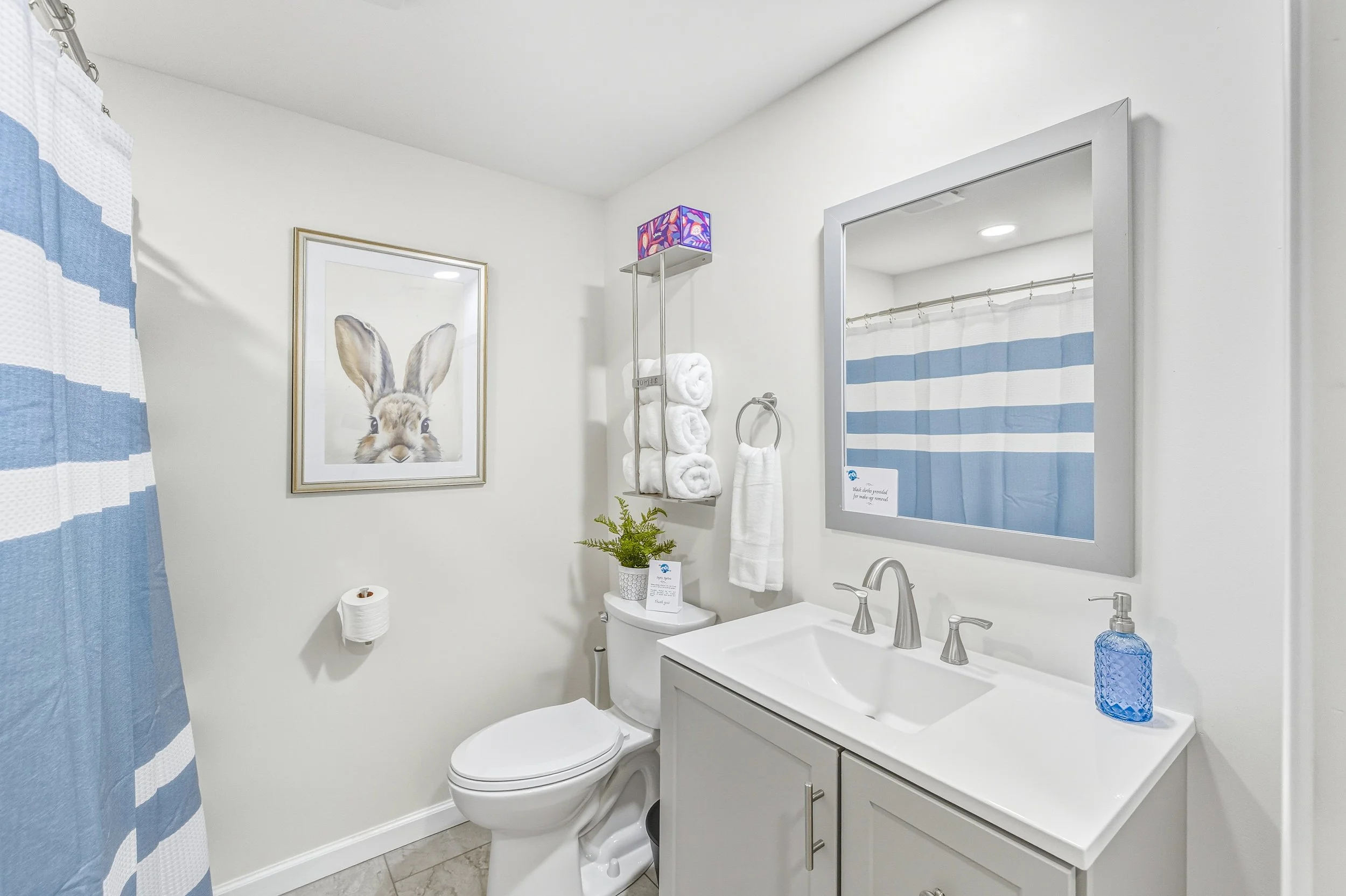 A bathroom with a white vanity and sink, a large mirror, white towels on a metal shelf, a bunny artwork on the wall, a purple lit lamp, a roll of toilet paper, and a blue textured soap dispenser on the sink. There is a blue and white striped shower c