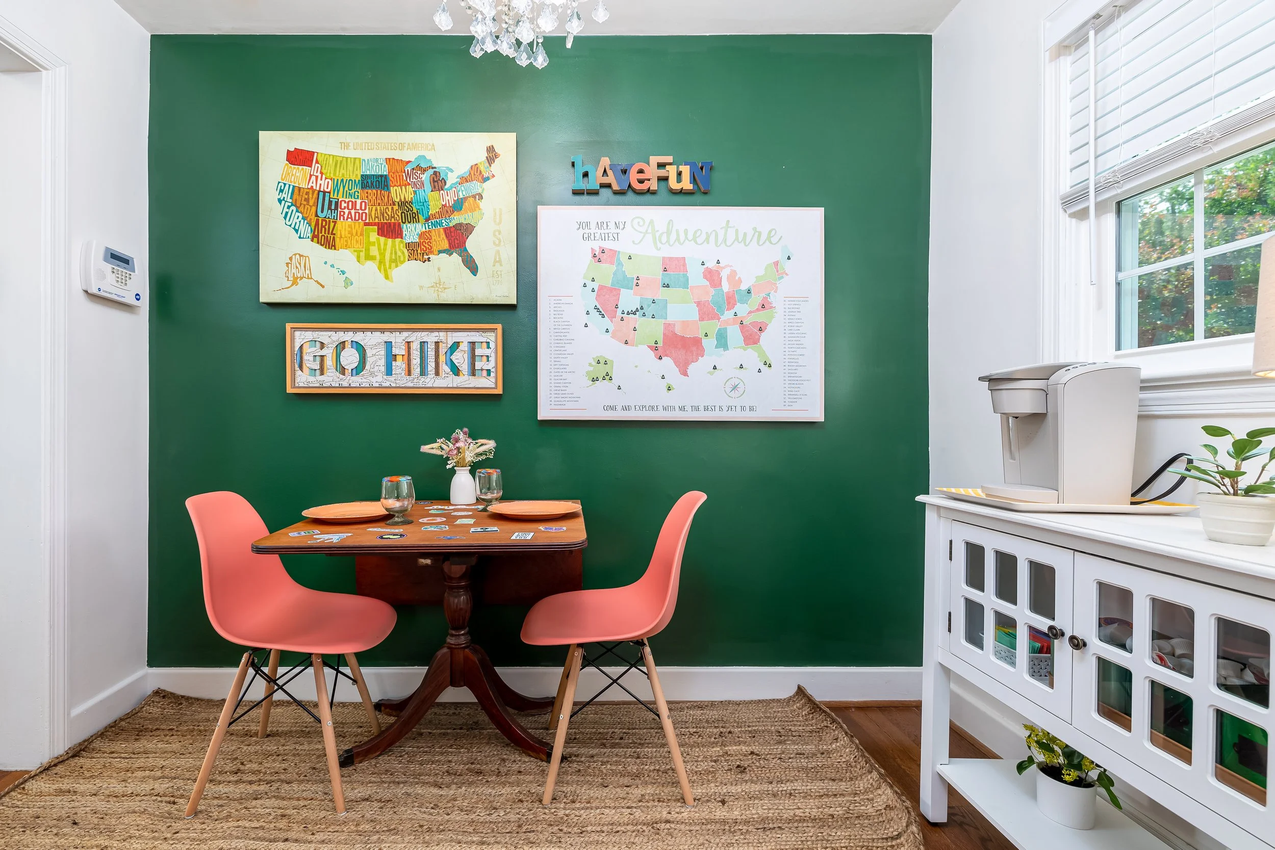 A dining room with a green accent wall decorated with maps and framed artwork, pink chairs, a wooden table, and white storage cabinet, near a window with blinds and greenery outside.