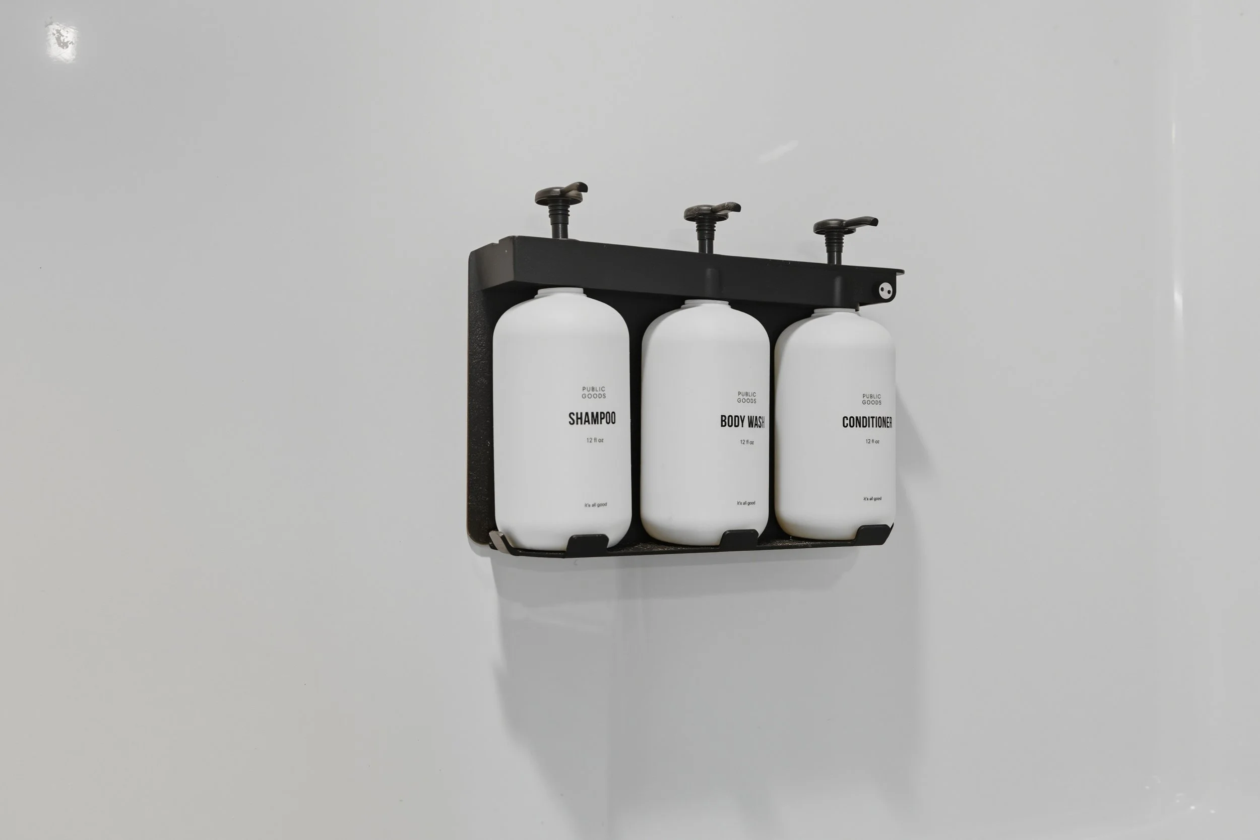 Three white bottles labeled 'SHAMPOO', 'BODY WASH', and 'CONDITIONER' are mounted on a black wall holder against a white wall.
