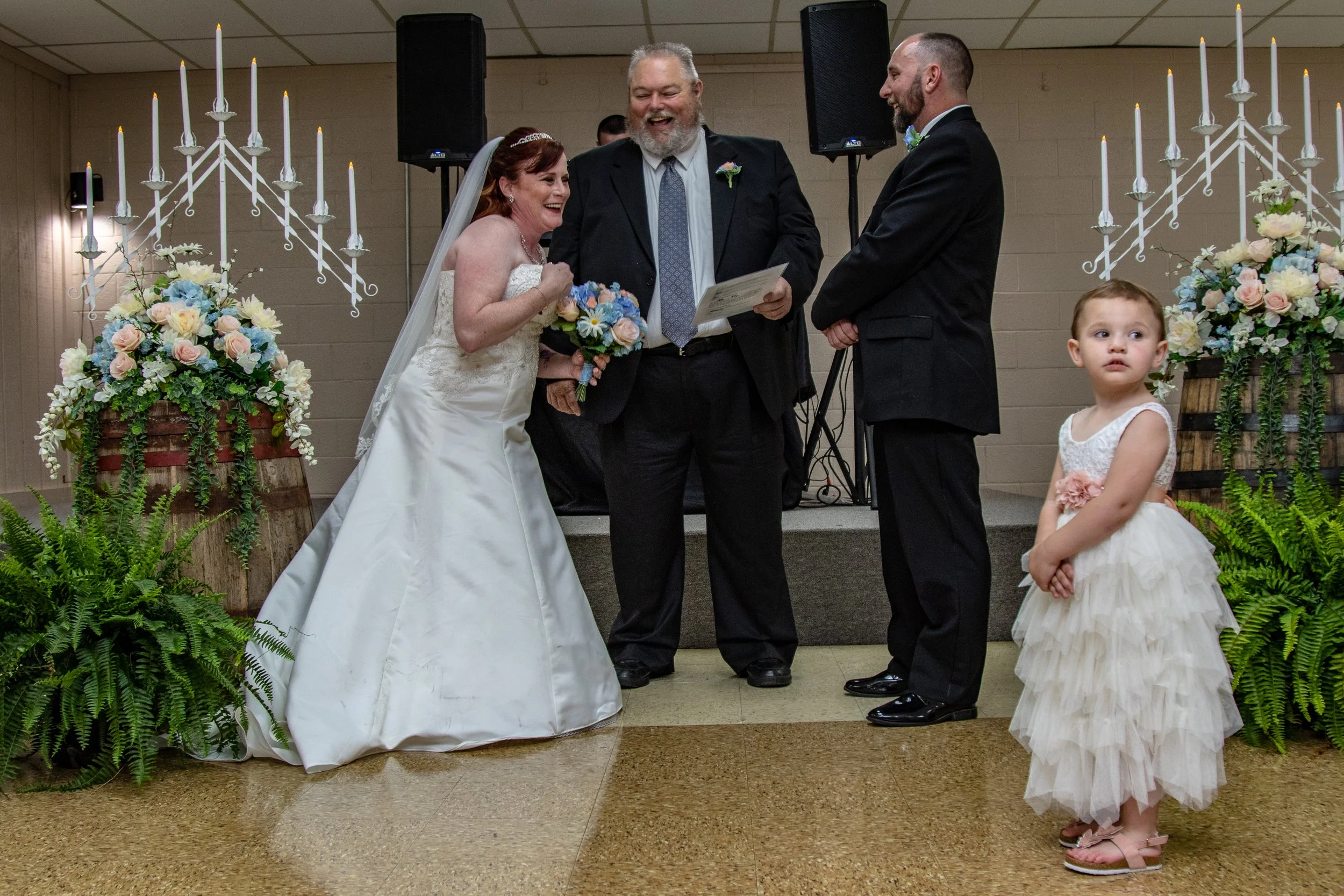 A wedding ceremony with a bride and groom standing before an officiant, surrounded by floral arrangements and candles, with a young girl dressed in a white dress standing nearby.