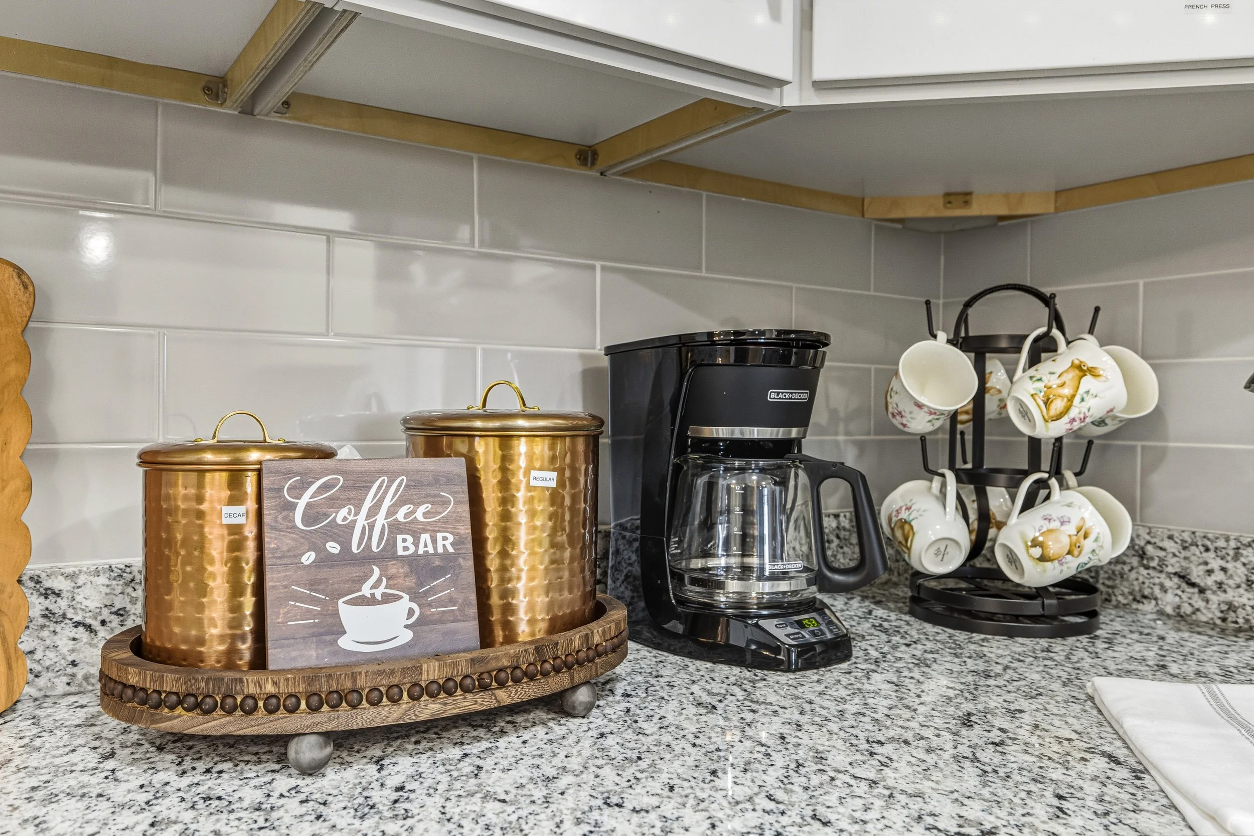 A kitchen countertop with a coffee bar setup, including three copper-colored jars labeled 'Decaf,' 'Regular,' and 'Dark Roast,' a decorative sign that reads 'Coffee Bar' with a coffee cup illustration, a Black & Decker coffee maker, a glass coffee po