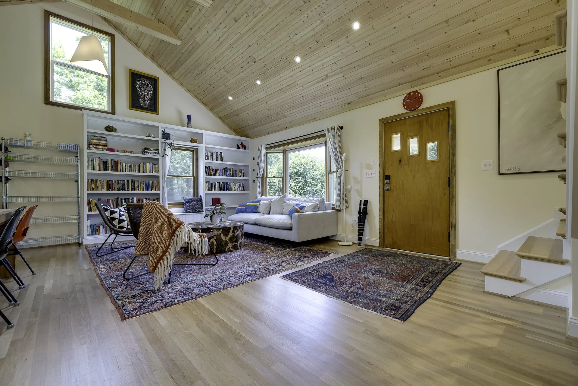Living room with wooden vaulted ceiling, large windows with white curtains, a white sofa with colorful pillows, a rug, a bookshelf filled with books, a red wall clock, and stairs leading upward.