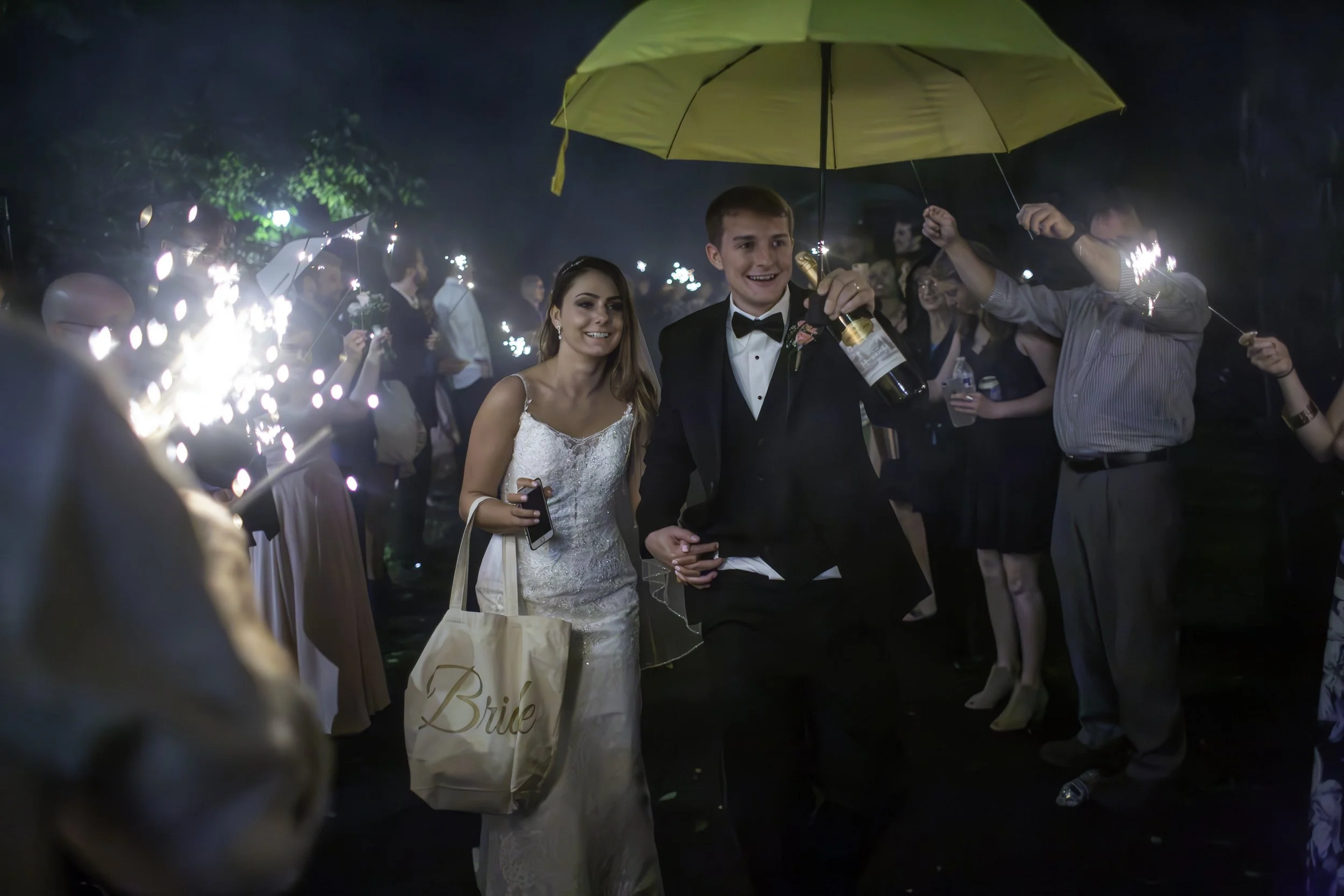 A newlywed couple at night holding a yellow umbrella, surrounded by guests celebrating with sparklers; the bride is wearing a white lace wedding dress, and the groom is in a black tuxedo with a bow tie, smiling happily.
