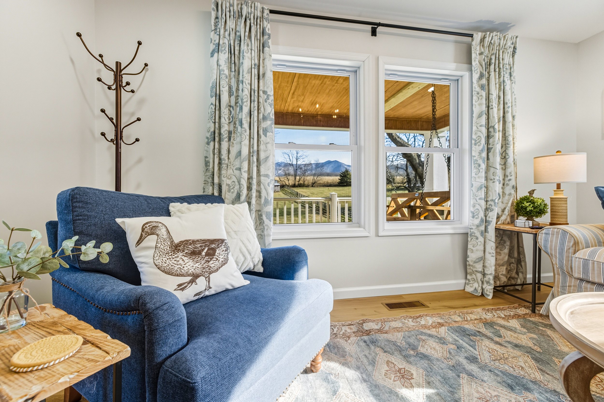 Living room with a blue armchair, decorative pillows, a wooden side table with yellow coasters, greenery, and a rug. Large windows with floral curtains reveal a porch with a swing and scenic mountain view.