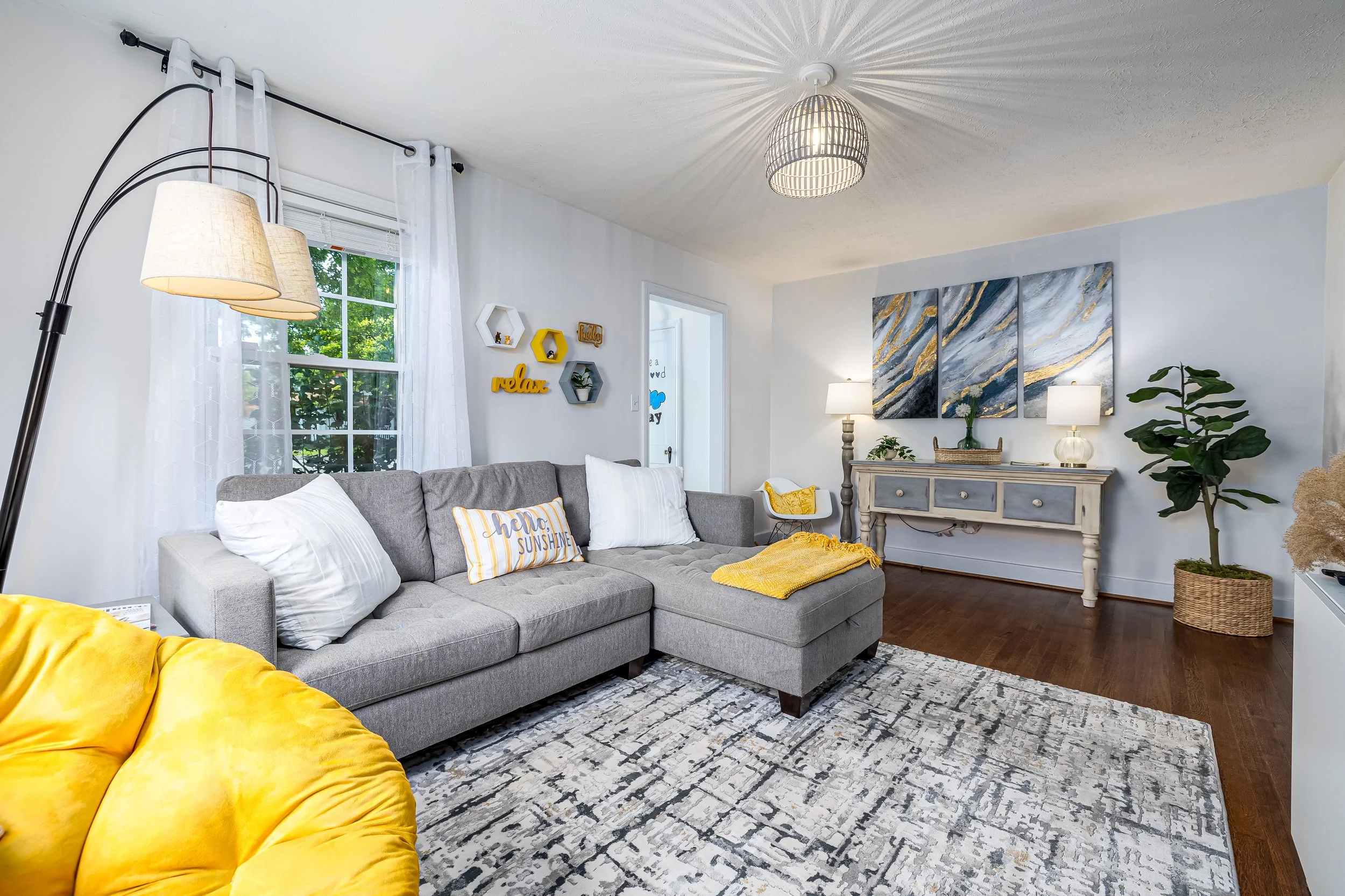 Living room with gray sectional sofa, yellow throw blanket, and pillows, large window with white curtains, wall decor including hexagonal shelves and a wooden sign that says 'relax,' modern art piece on wall, two table lamps on a sideboard, potted pl