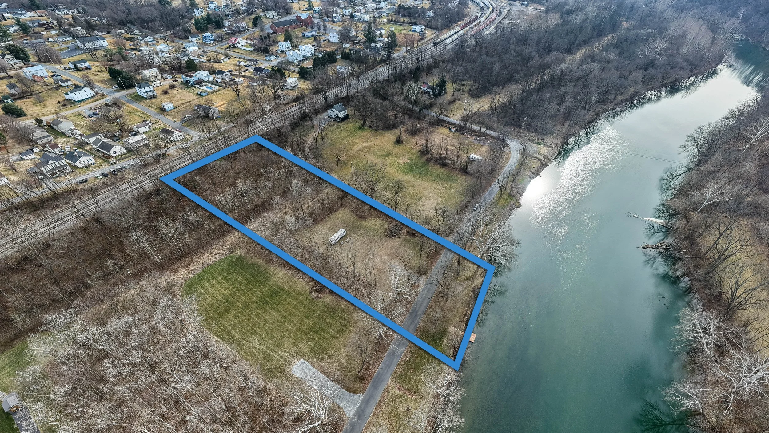 Aerial view of a rectangular plot of land marked with a blue outline, next to a body of water, with trees, a small building, and grassy areas, in a suburban neighborhood.